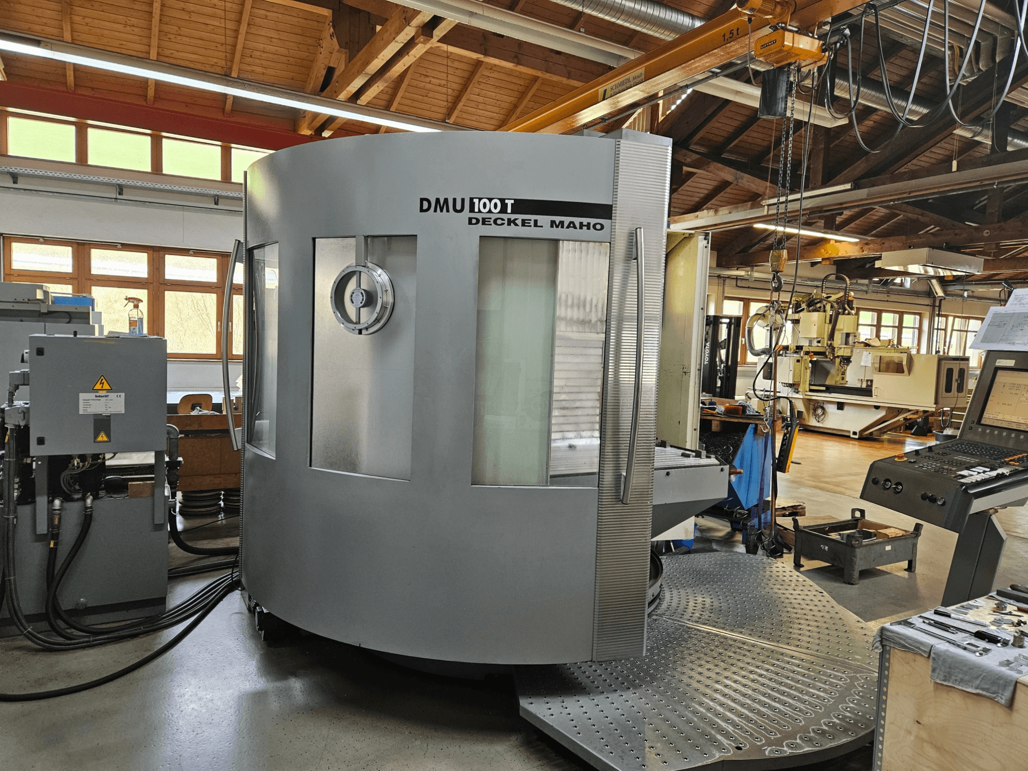 DMU 100 T Deckel Maho CNC-tööpink, mille esiplaanil on suur ümmargune alus, juhtpaneel ja klaasist korpus.
