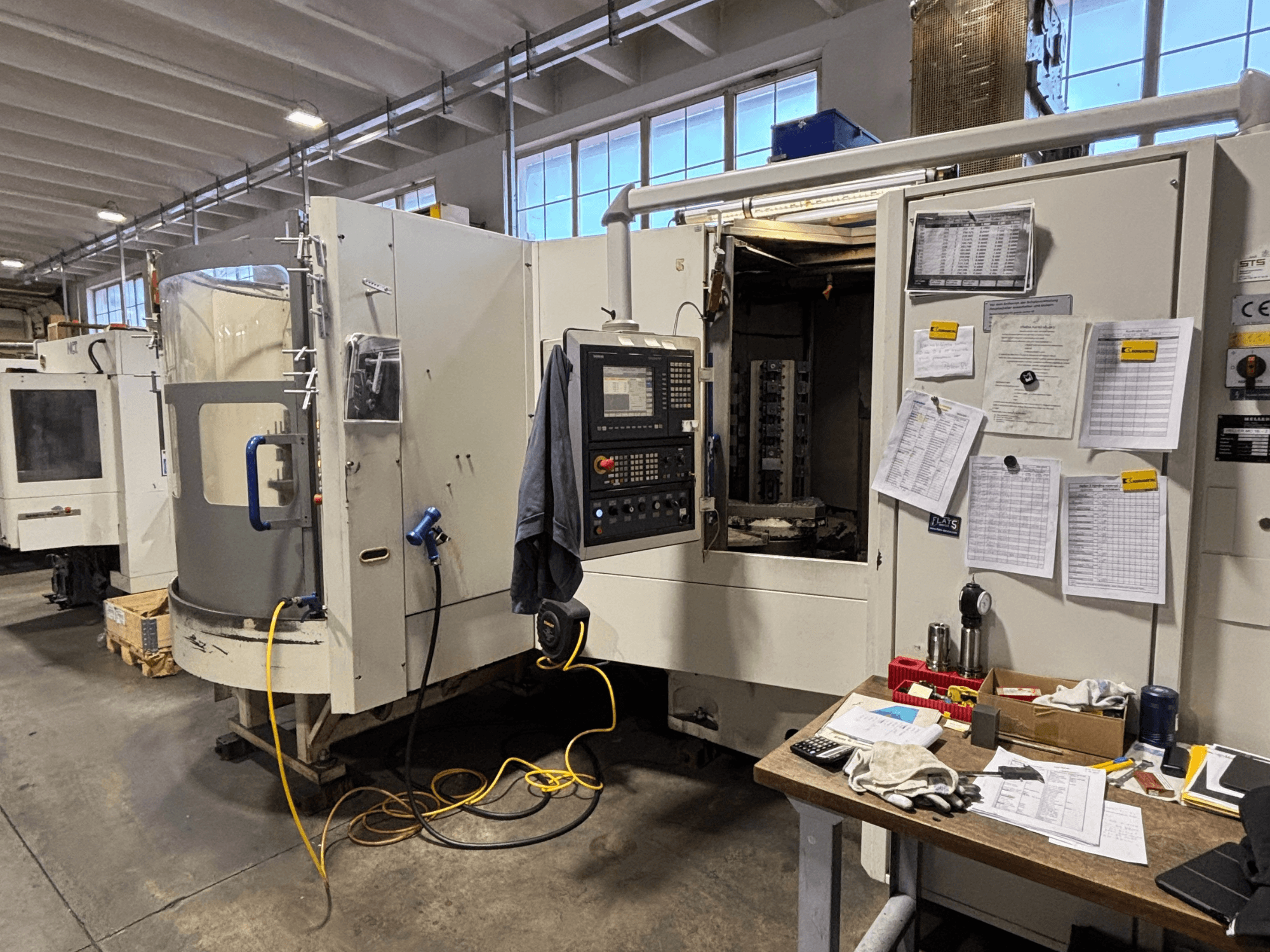 CNC-tööpink töökojas, esiplaanil, küljel juhtpaneel ja dokumentatsioon; tööriistad ja materjalid laual.