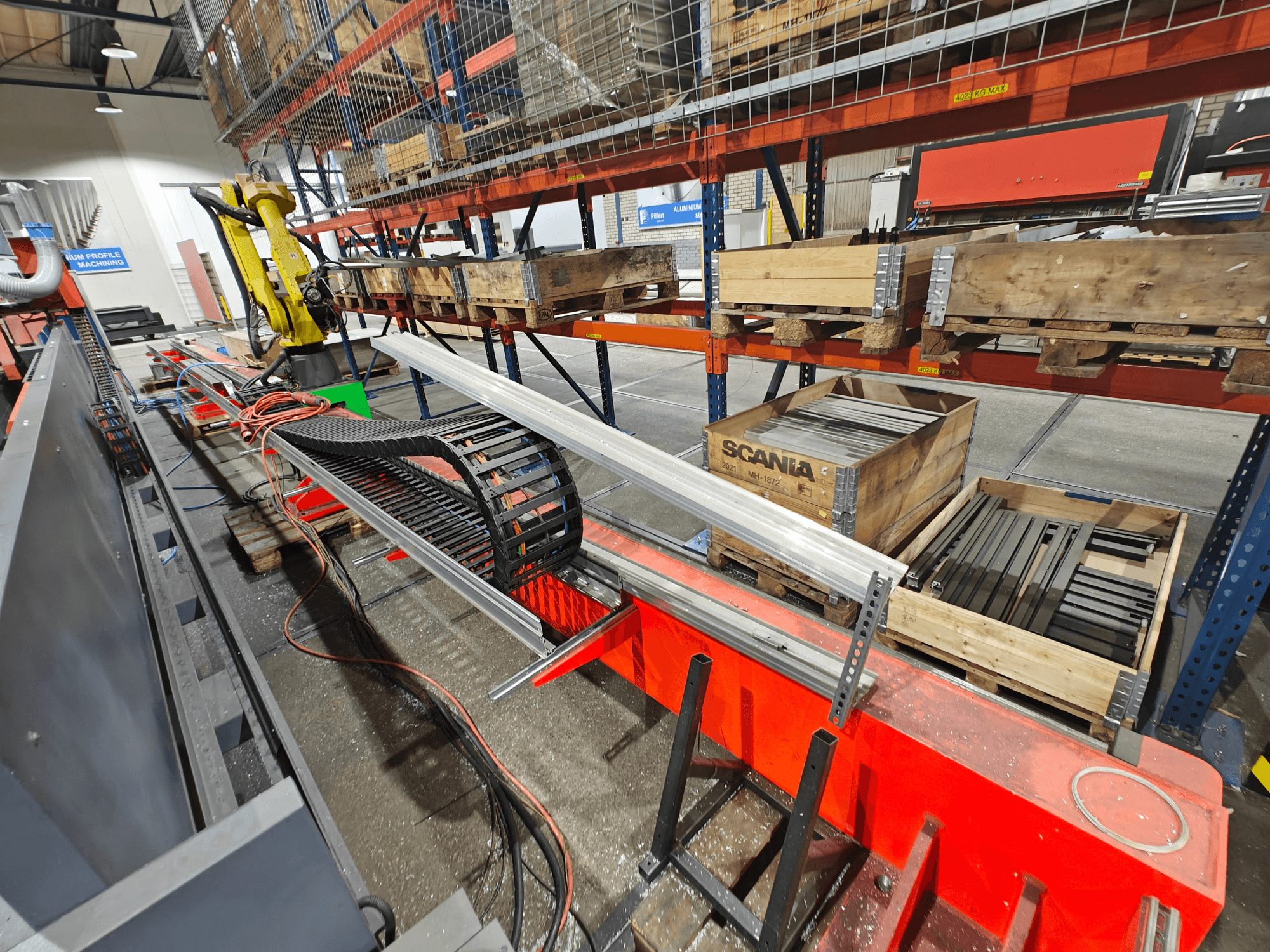 Robotkäsi tööjaamas, oranžid ja mustad kaablid nähtavad, koos Scania kastide ja alumiiniumprofiilidega tehases.