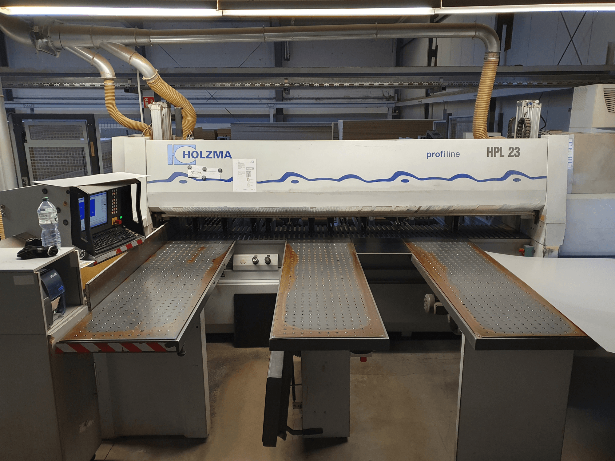 HOLZMA HPL 23 lõikemasin esiplaanil, kolm perforeeritud lauda, arvutiterminal ja veepudel lähedal.