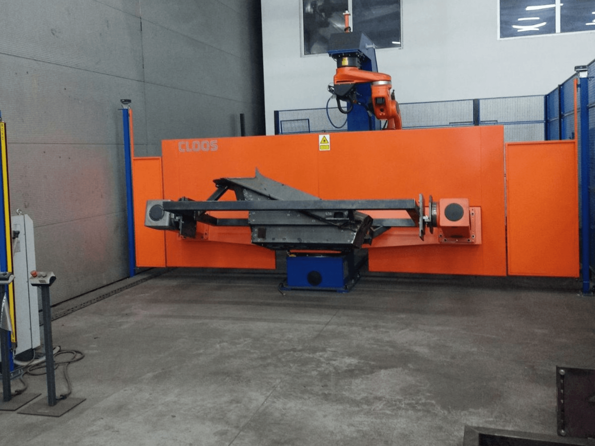 CLOOS QIROX QRC-350-E automatiseerimissüsteemi esiplaanil, robotkäe ja metallkonstruktsiooniga tööstuslikus keskkonnas.