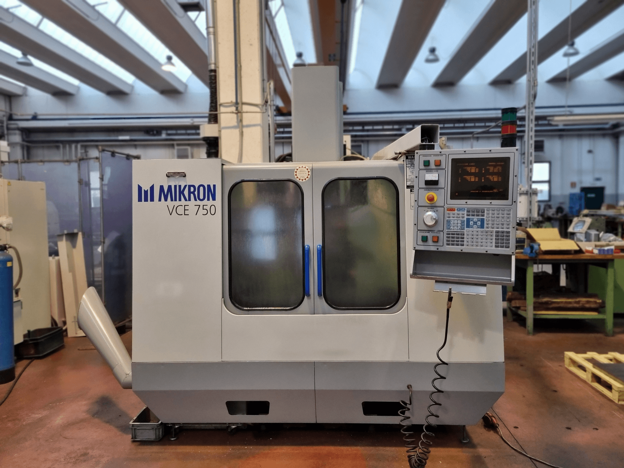 Valge Mikron VCE 750 CNC-tööpink töökojas, külgvaade, digitaalse juhtpaneeli ja suurte klaasist ustega.