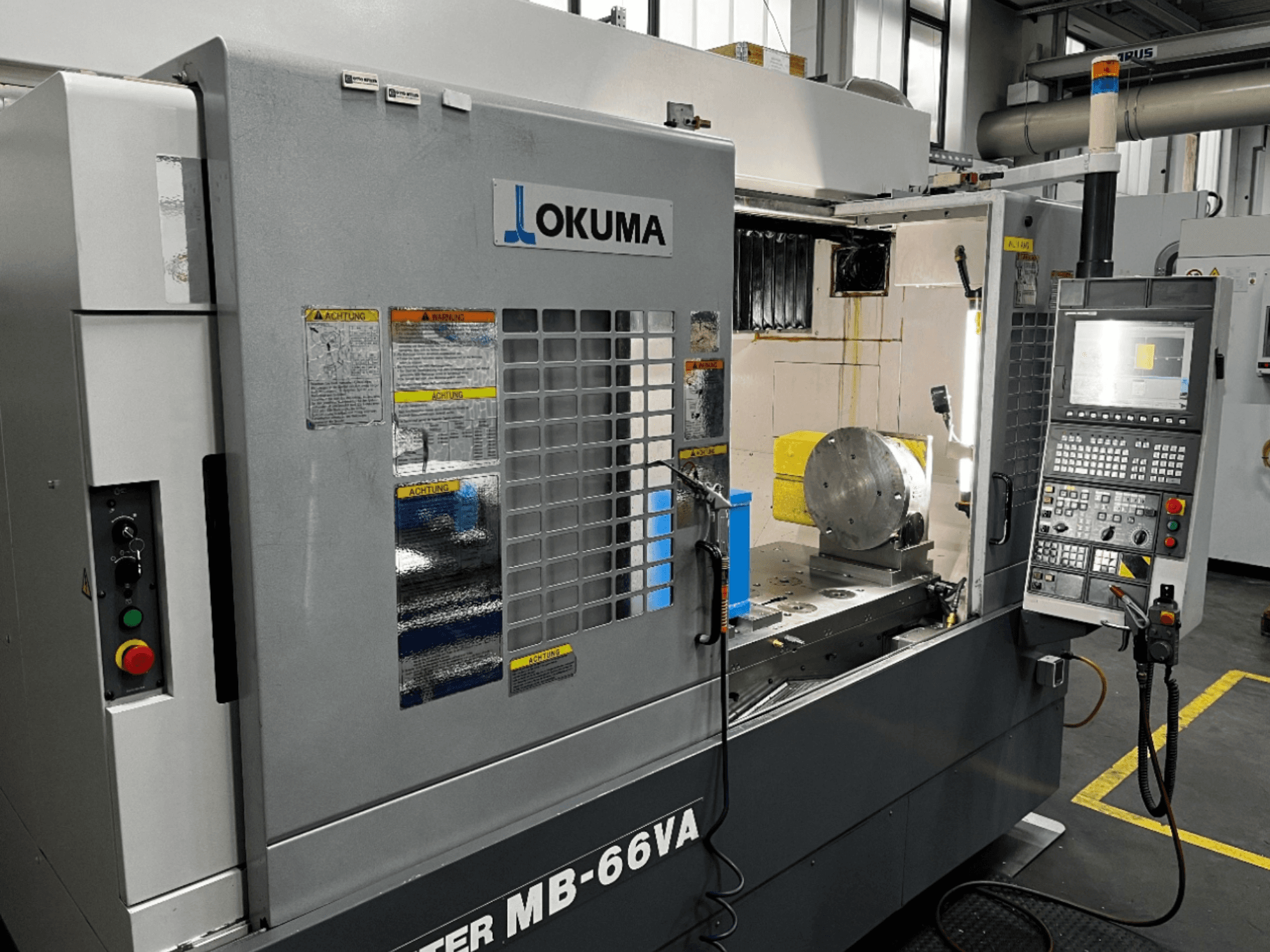 Okuma MB66-VA vertikaalne töötlemiskeskus esiplaanil, kus on näidatud juhtpaneel, töödeldav detail ja masina sisemus koos ohutussiltidega.