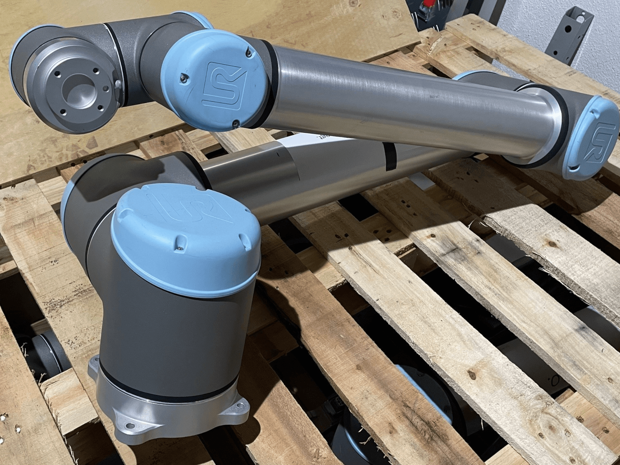 Hõbedane robotkäsi siniste otsakattega, esiplaanil, näidates liigeseid ja mehaanilisi detaile, toetub puidust kaubaalusele.