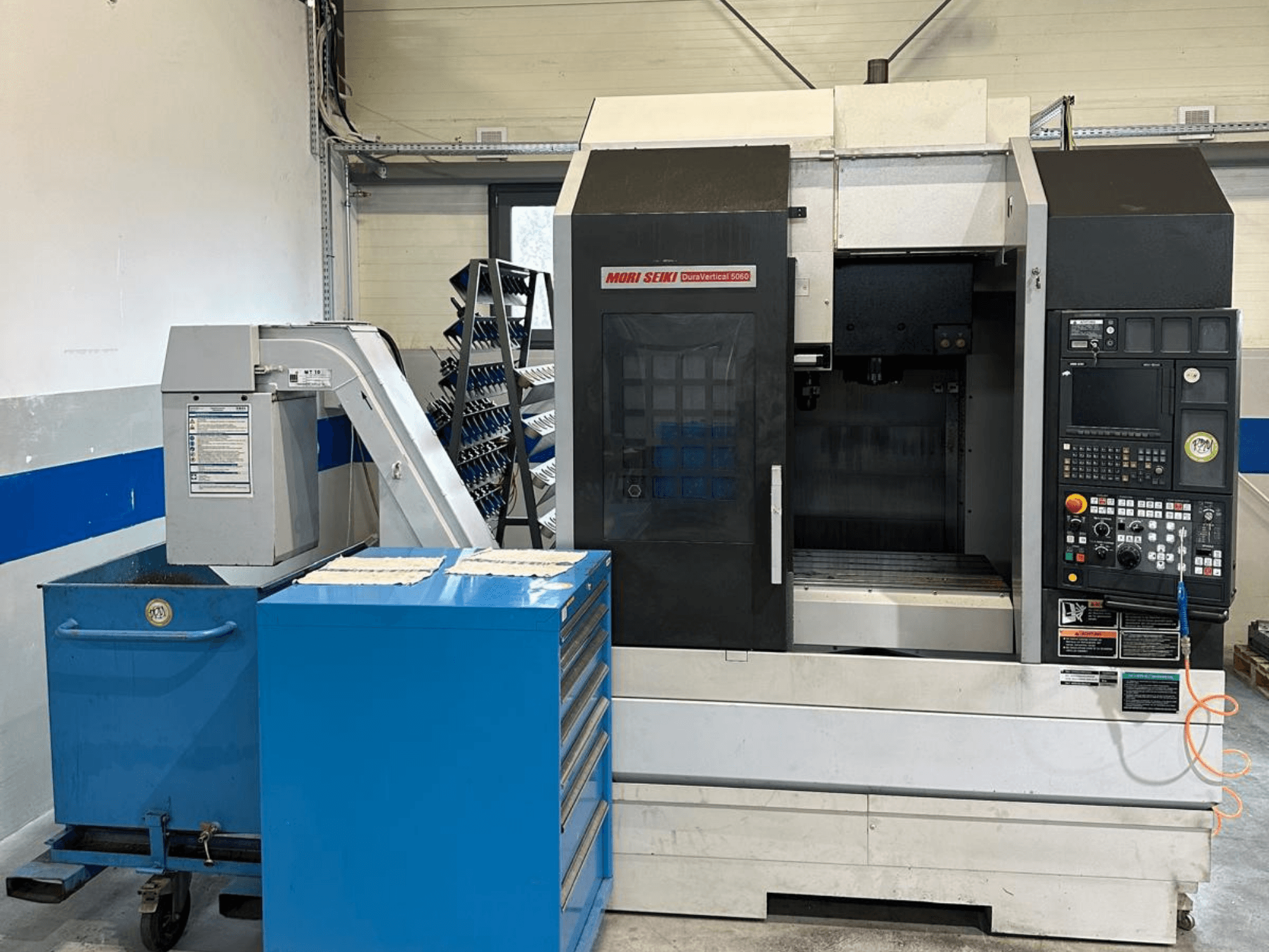 MORI SEIKI DURA VERTICAL 5060 – vertikaalse töötlemiskeskuse esivaade koos juhtpaneeli ja selle lähedal asuva tööriistade hoiukohaga.