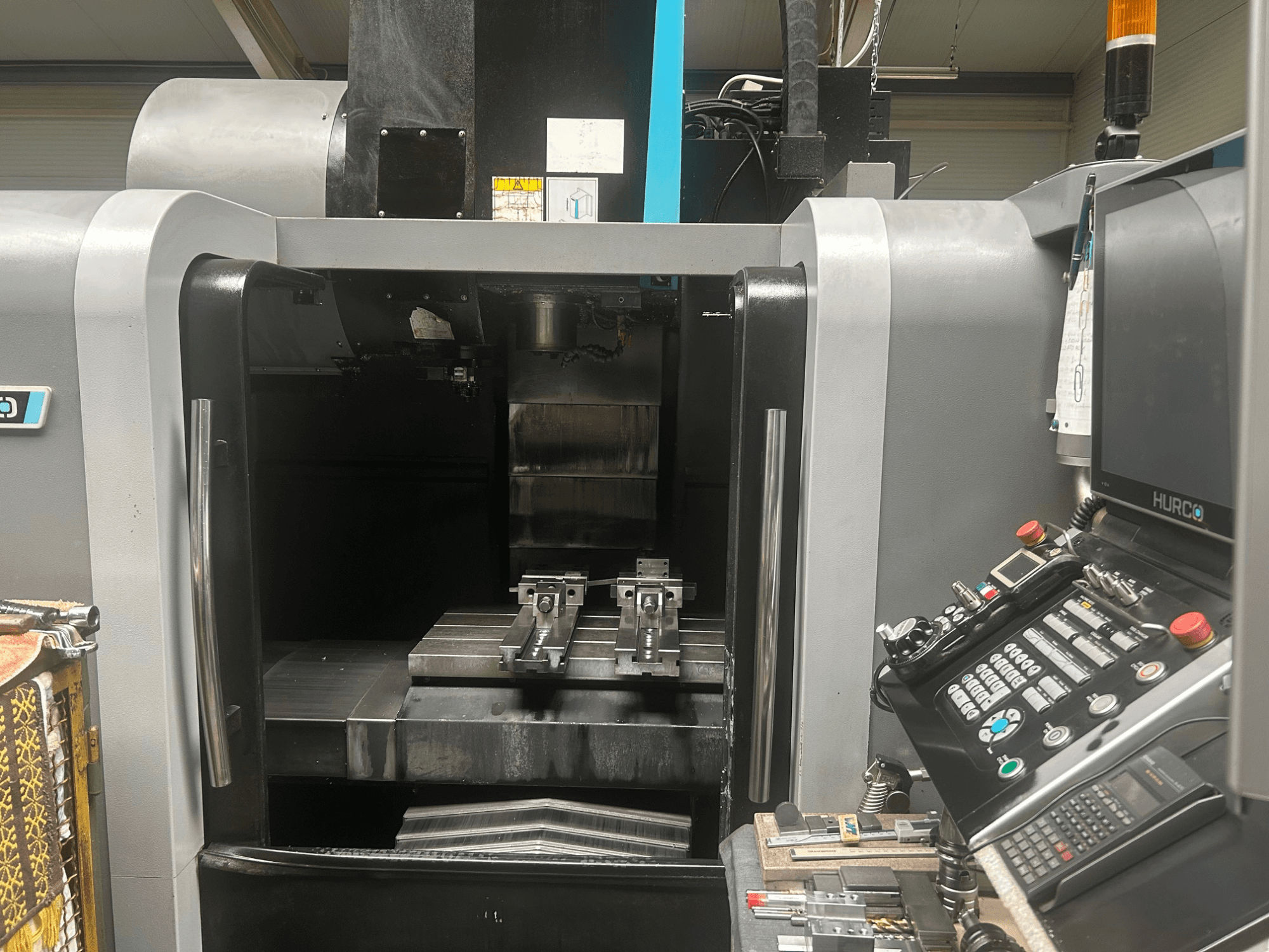 Hurco CNC-töötluskeskuse esivaade, millel on kujutatud tööriistahoidja, metalltöödeldis ning juhtpaneel koos nuppude ja ekraaniga.