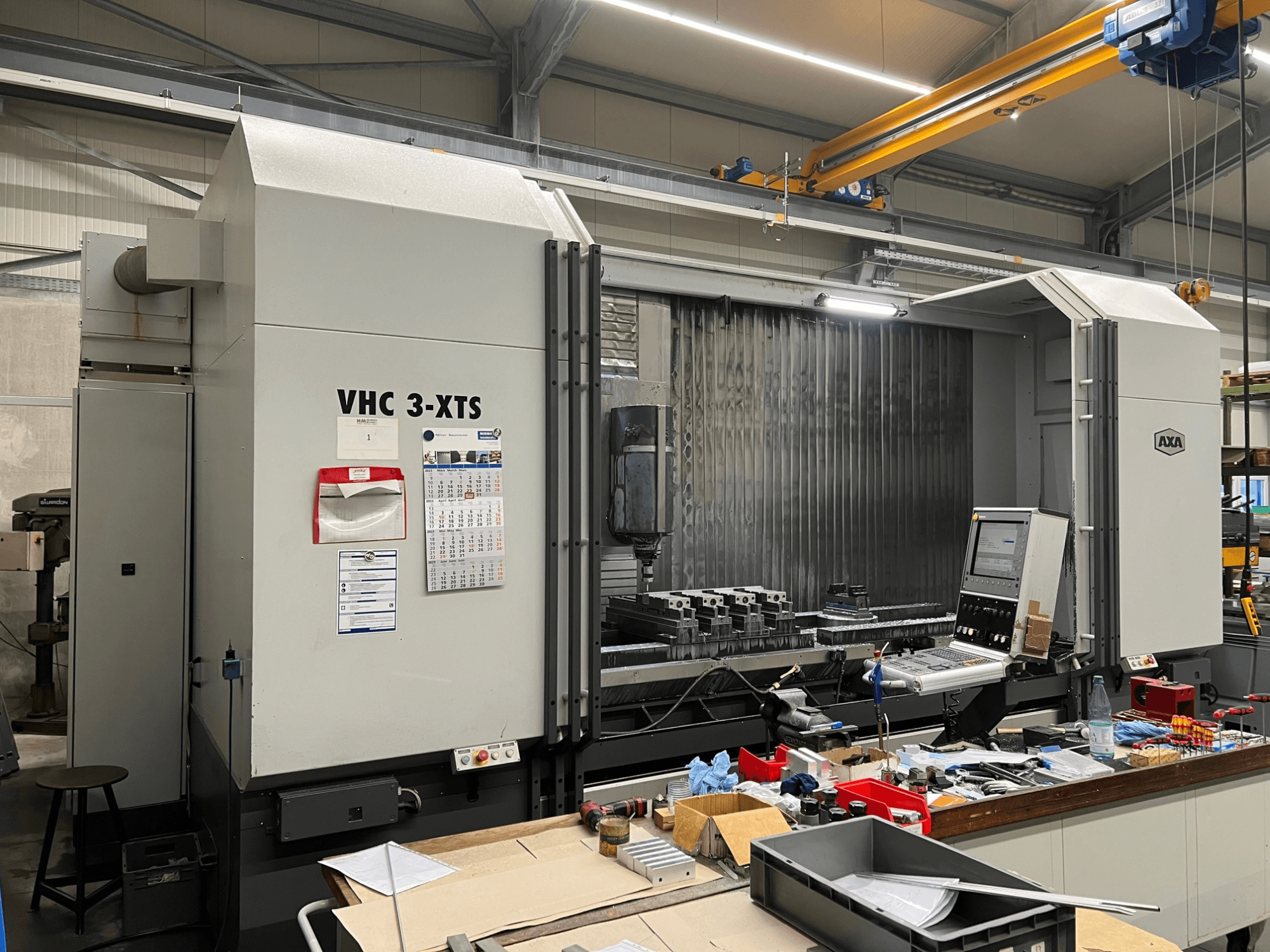 AXA VHC 3-XTS CNC-tööpingi külgvaade, juhtpaneel ja tööala täis tööriistu ja komponente.
