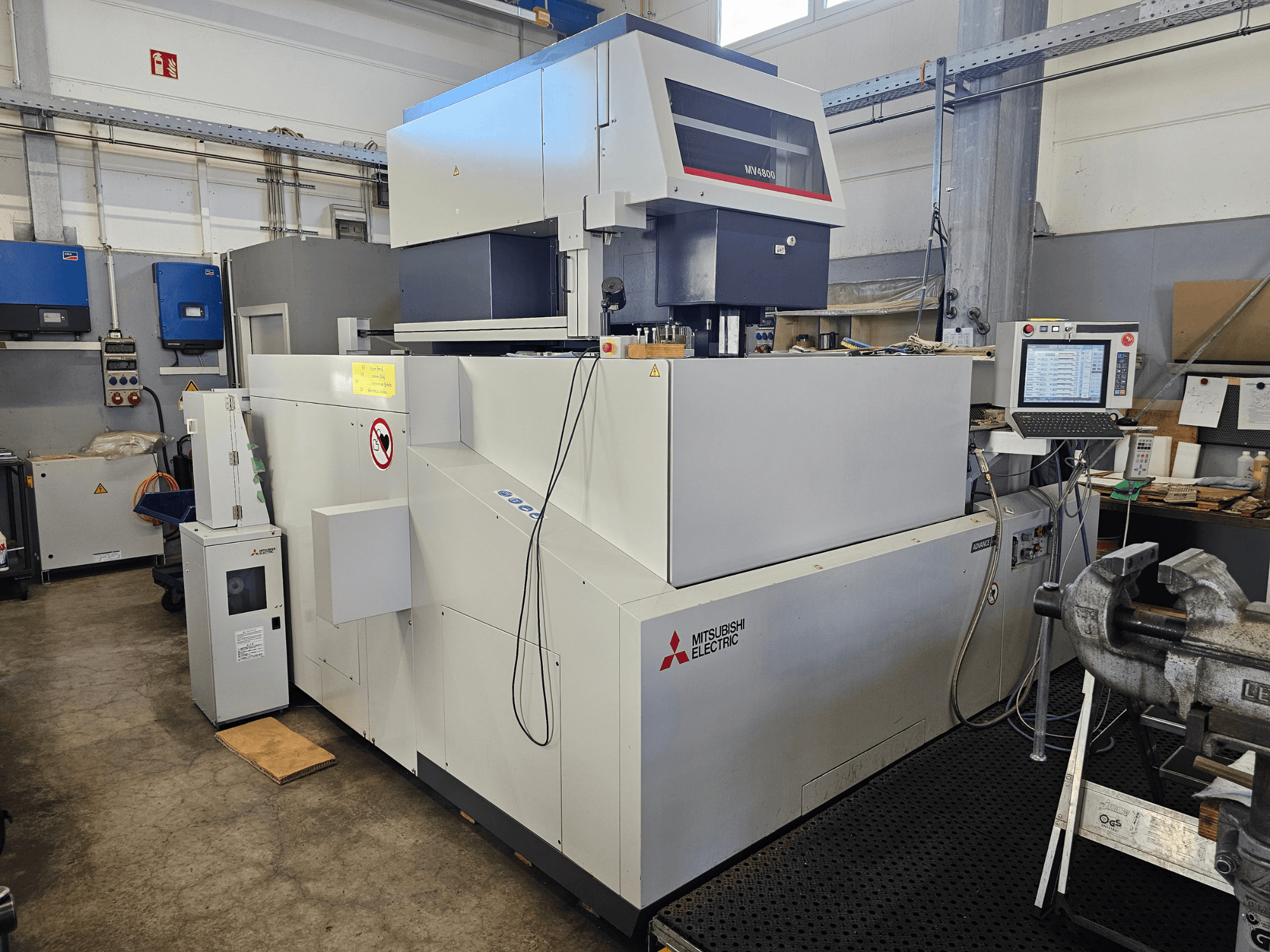 Mitsubishi MV4800 traadikeeramismasin töökojas, külgvaates on näha juhtpaneel, ohutussildid ja ümbritsevad seadmed.