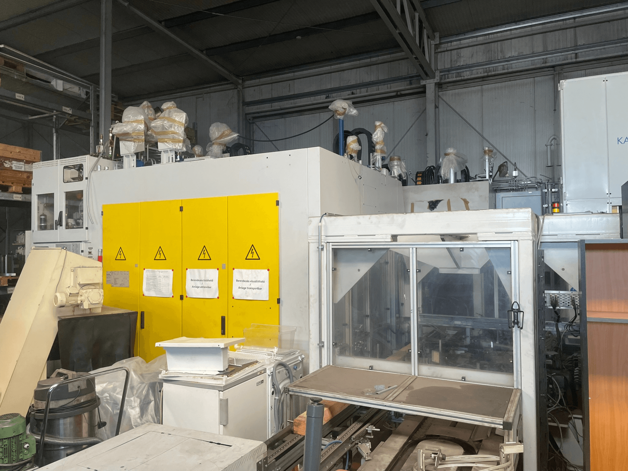 Kollase juhtpaneeliga tööstusmasinad, külgvaates, hoiatussiltide ja plastkattega, laoümbruses.