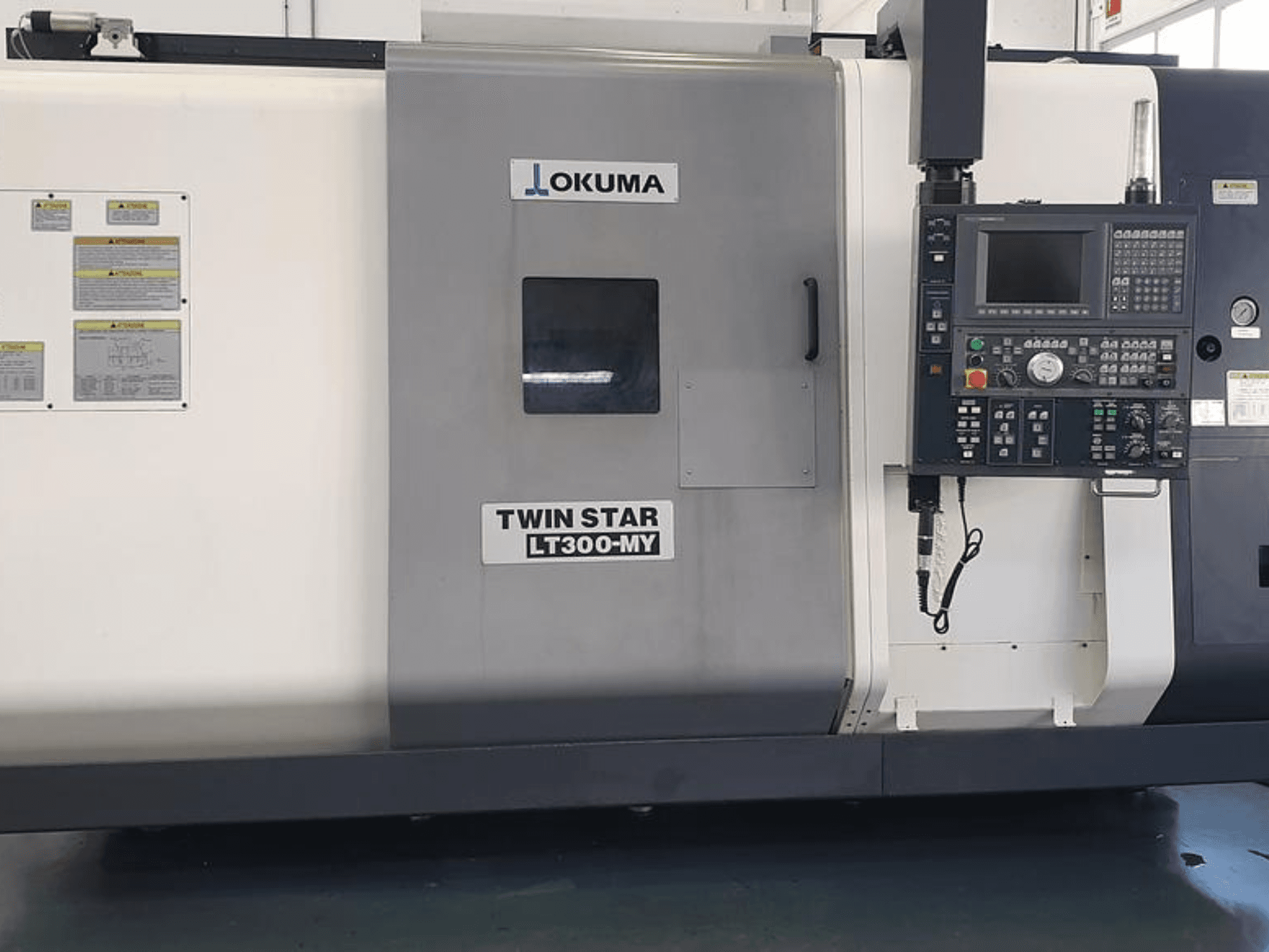 Okuma Twin Star LT300-MY CNC-teeninduspingi külgvaade, millel on juhtpaneel, juhtnupud ja ohutusmärgised.