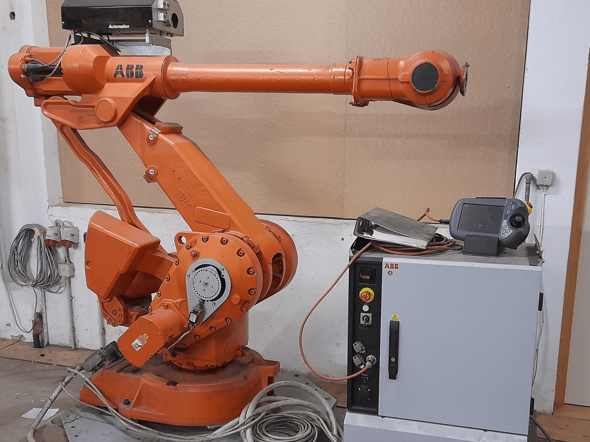 Oranž ABB robotkäsi koos juhtpaneeliga, esiplaanil, kus on näha kaablid ja automaatika seadmed.