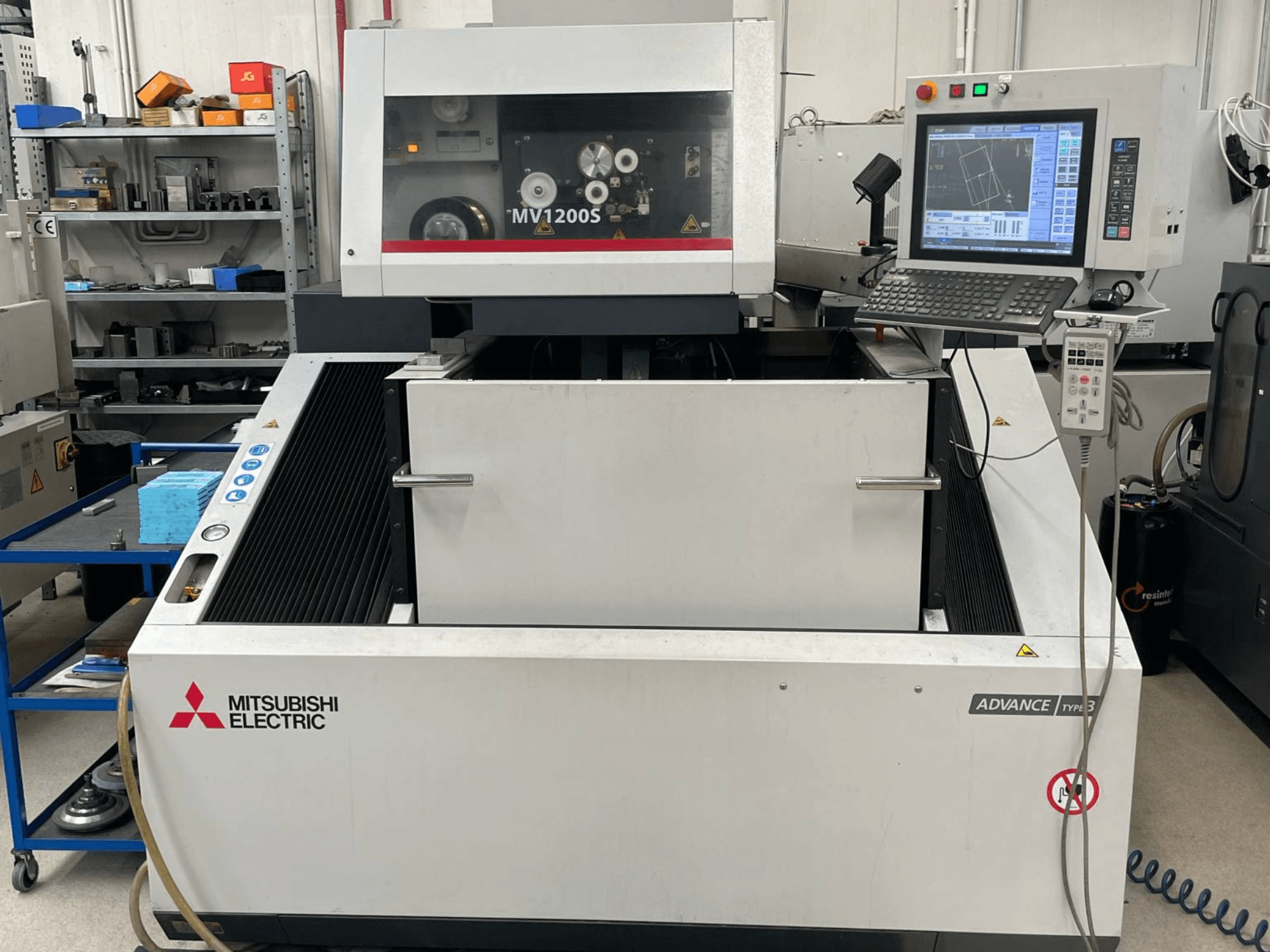 Mitsubishi MV1200S traat-EDM-masin eestvaates, millel on näha juhtpaneel ja tööpiirkond ning ohutushoiatused.