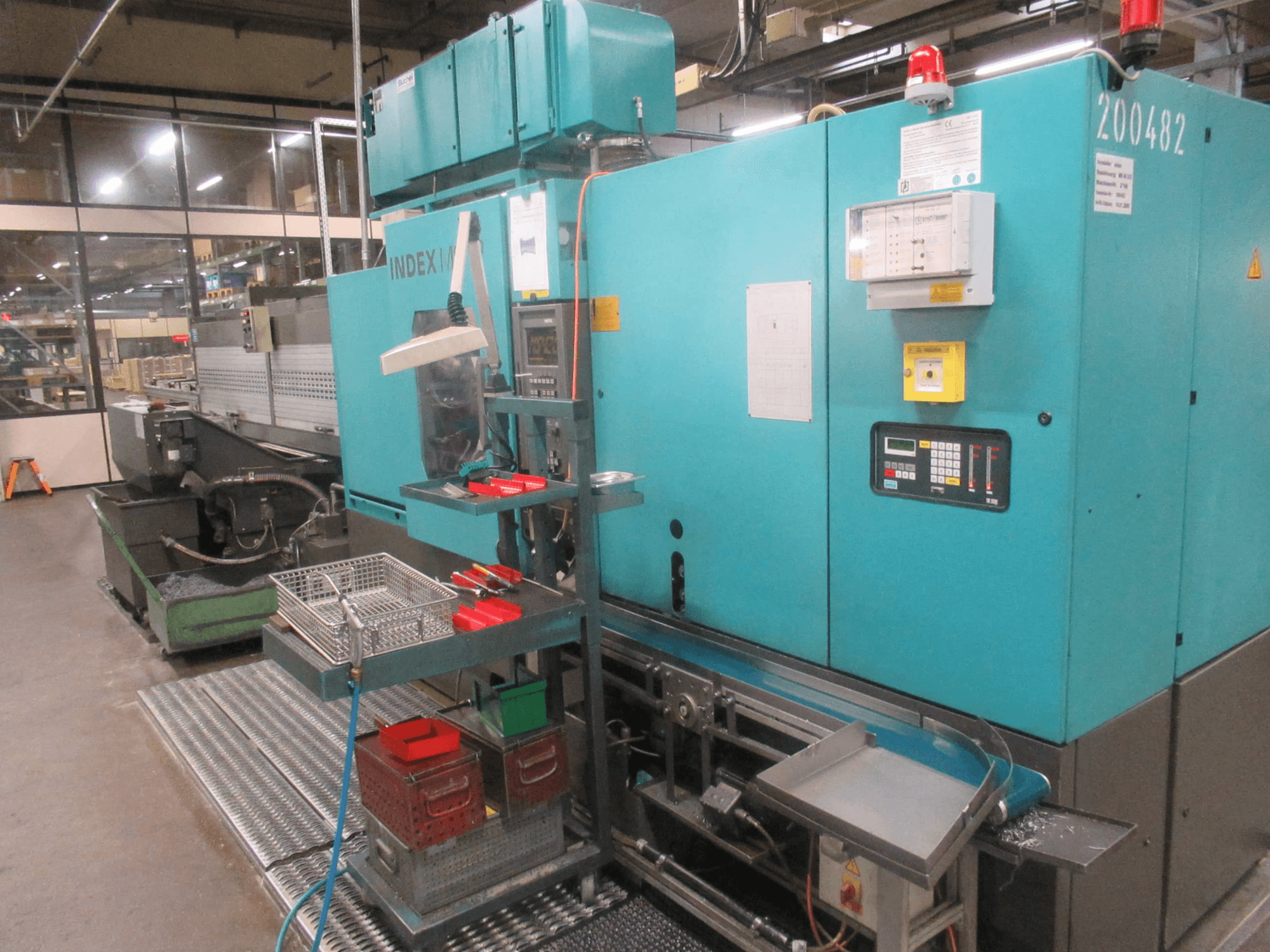 Türkiissinine INDEX masin tehases, külgvaade; juhtpaneel, tööriistade hoidla ja konveier. Nähtavad mahutid, voolikud ja häiresignaalid.