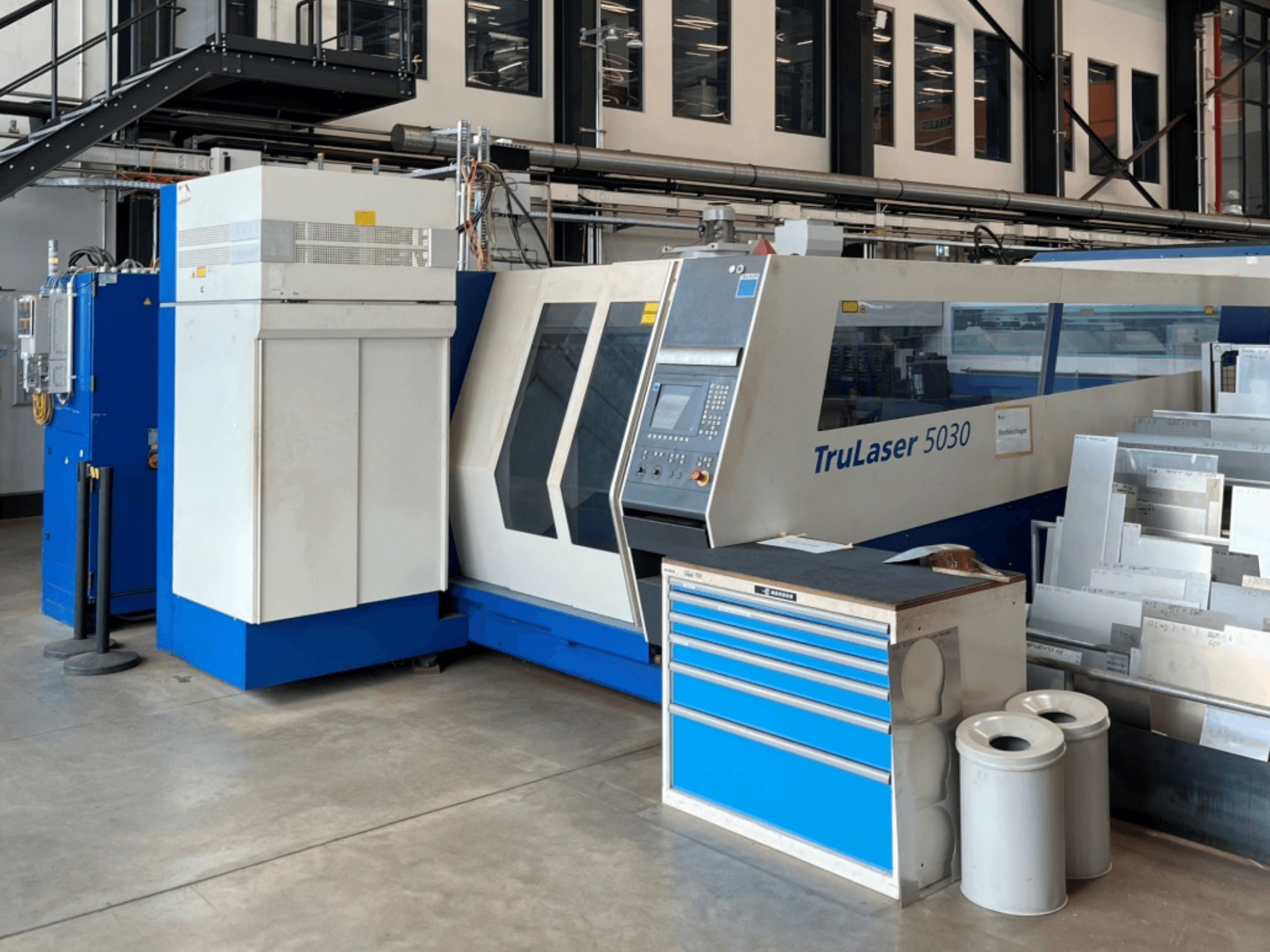 TRUMPF TruLaser 5030 CO₂-laserlõikur vaates eest, millel on juhtpaneel ja selle kõrval asuv töölaud metalllehtedega.