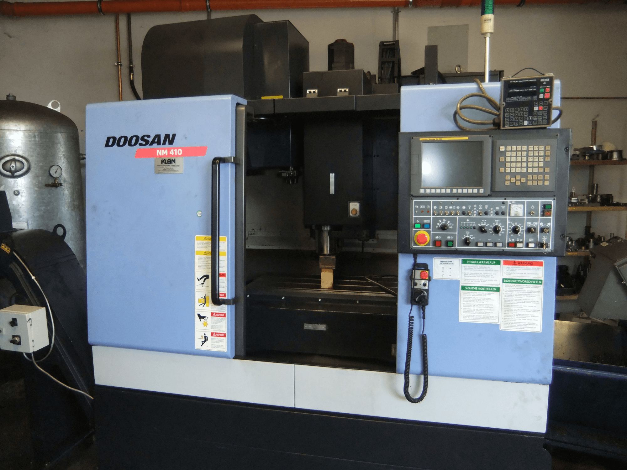 DOOSAN NM 410 vertikaalne töötlemiskeskus, vaade eest, millel on näha juhtpaneel, spindli piirkond ja ohutusmärgised.