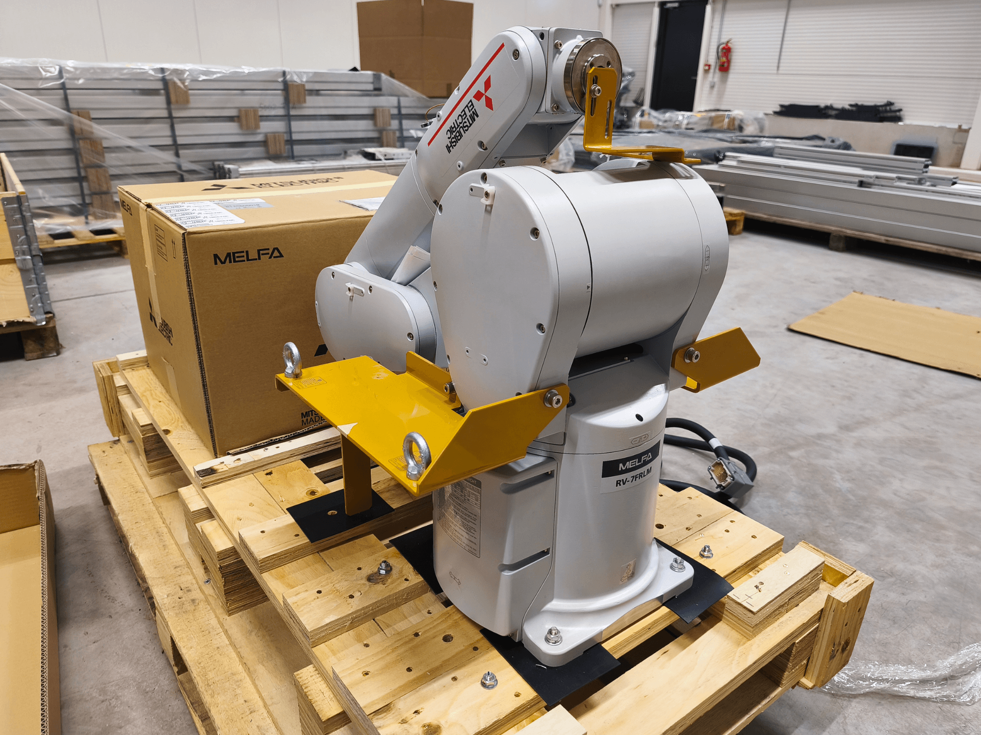 MELFA RV-7FLM tööstuslik robotkäsi puidust kaubaalusel, esiplaanil, kollaste tugikonksude ja halli pakendikarbiga.