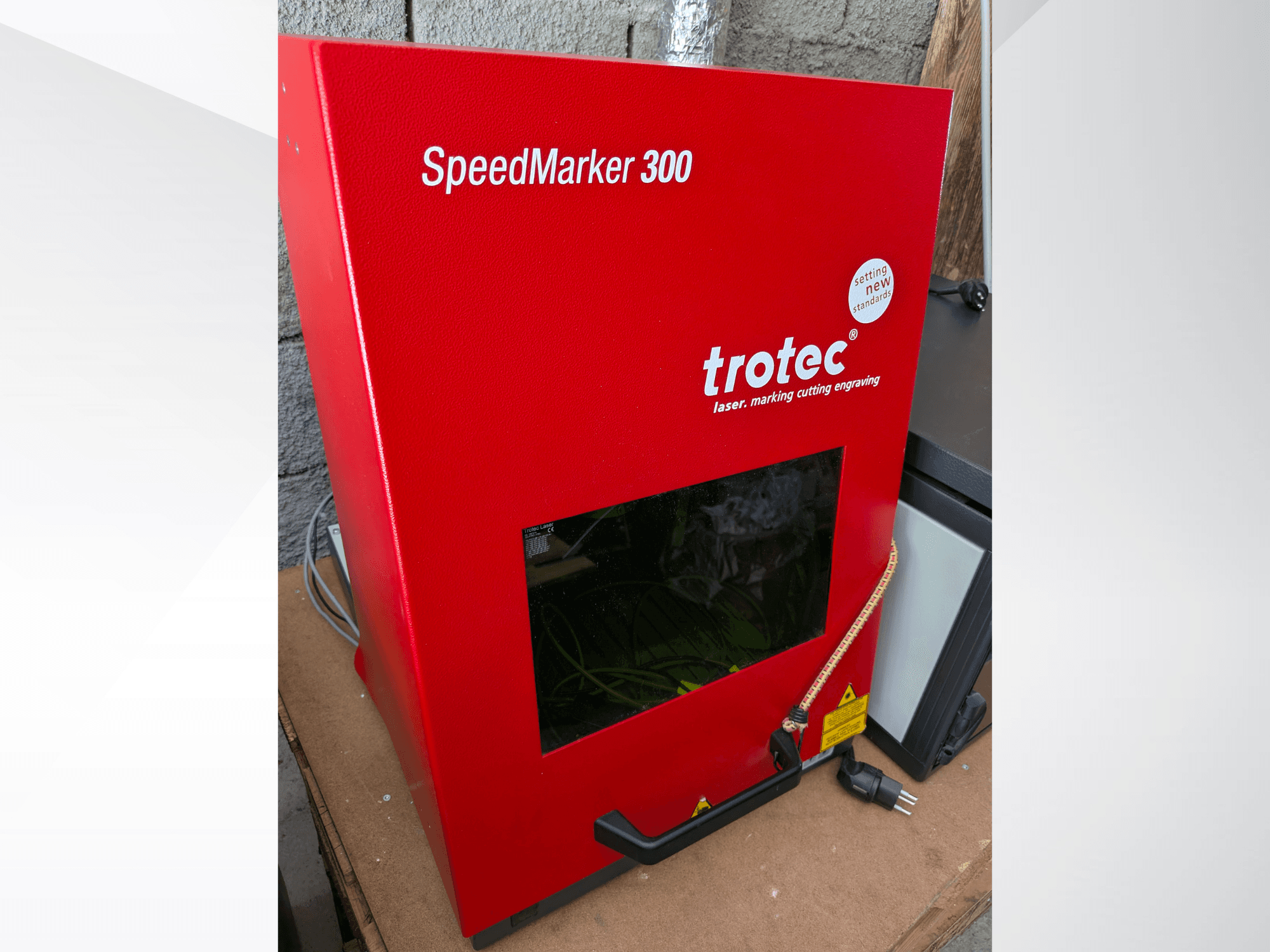 Punane Trotec SpeedMarker 300 lasermärgistusmasin esiplaanil, millel on suur vaateaken ja vooluühendus.