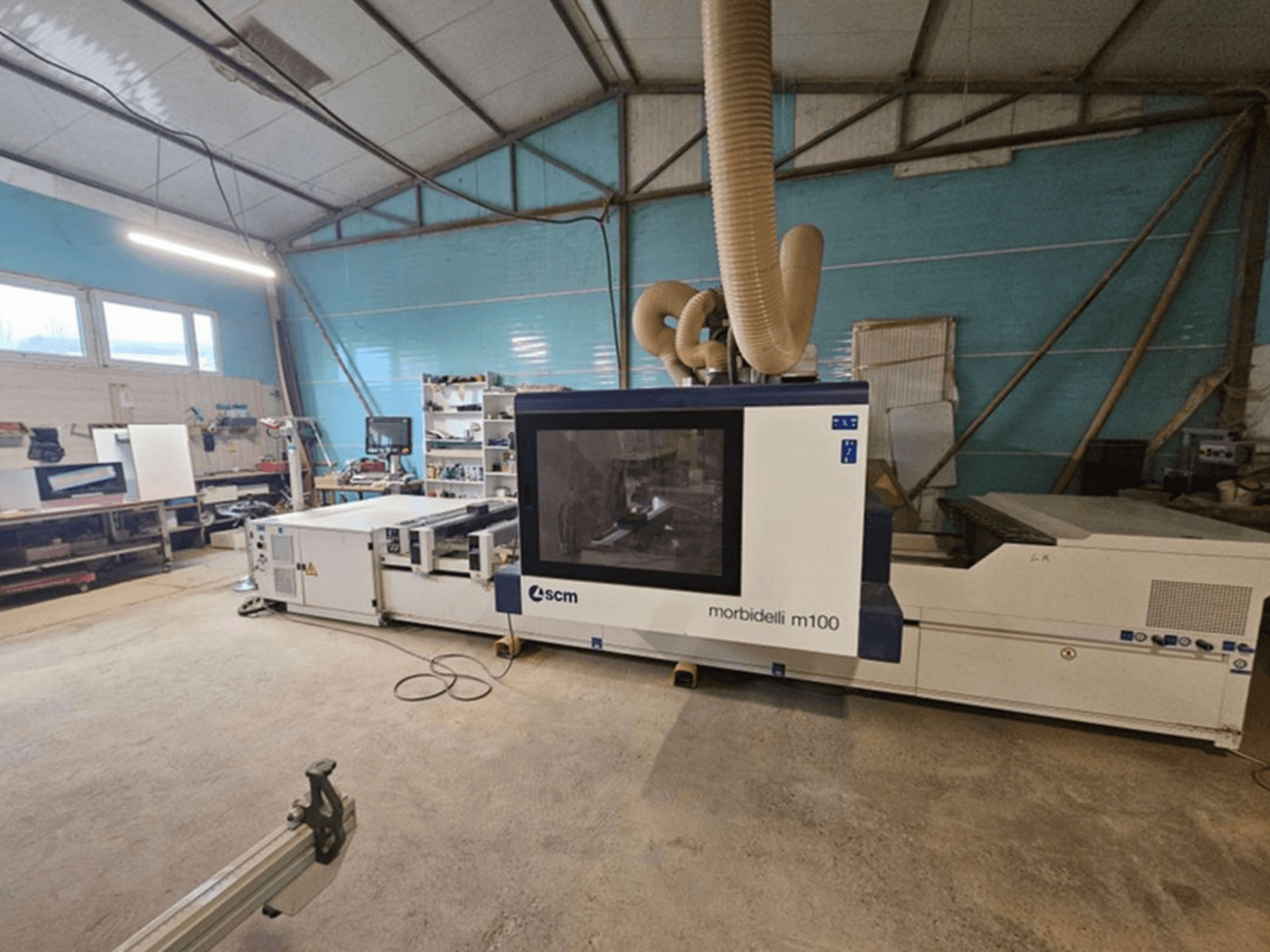 Töökojas asuv SCM MORBIDELLI M 100 CNC-töötlemiskeskus, vaade eest, koos tolmuärastussüsteemi ja ümbritseva töökoha varustusega.