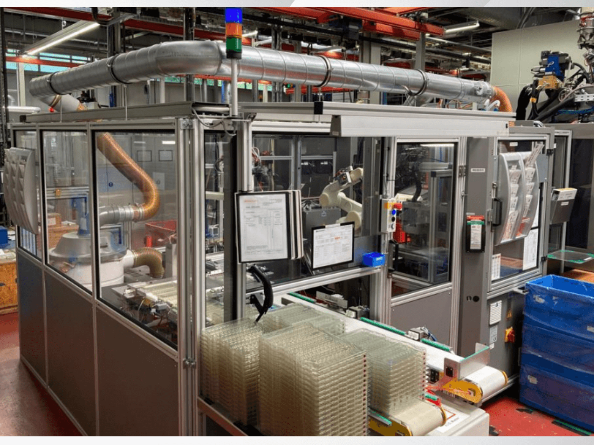 Eesvaade automatiseeritud masinale, millel on metalltorud, juhtpaneel ja virnastatud läbipaistvad plastkonteinerid tehase põrandal.