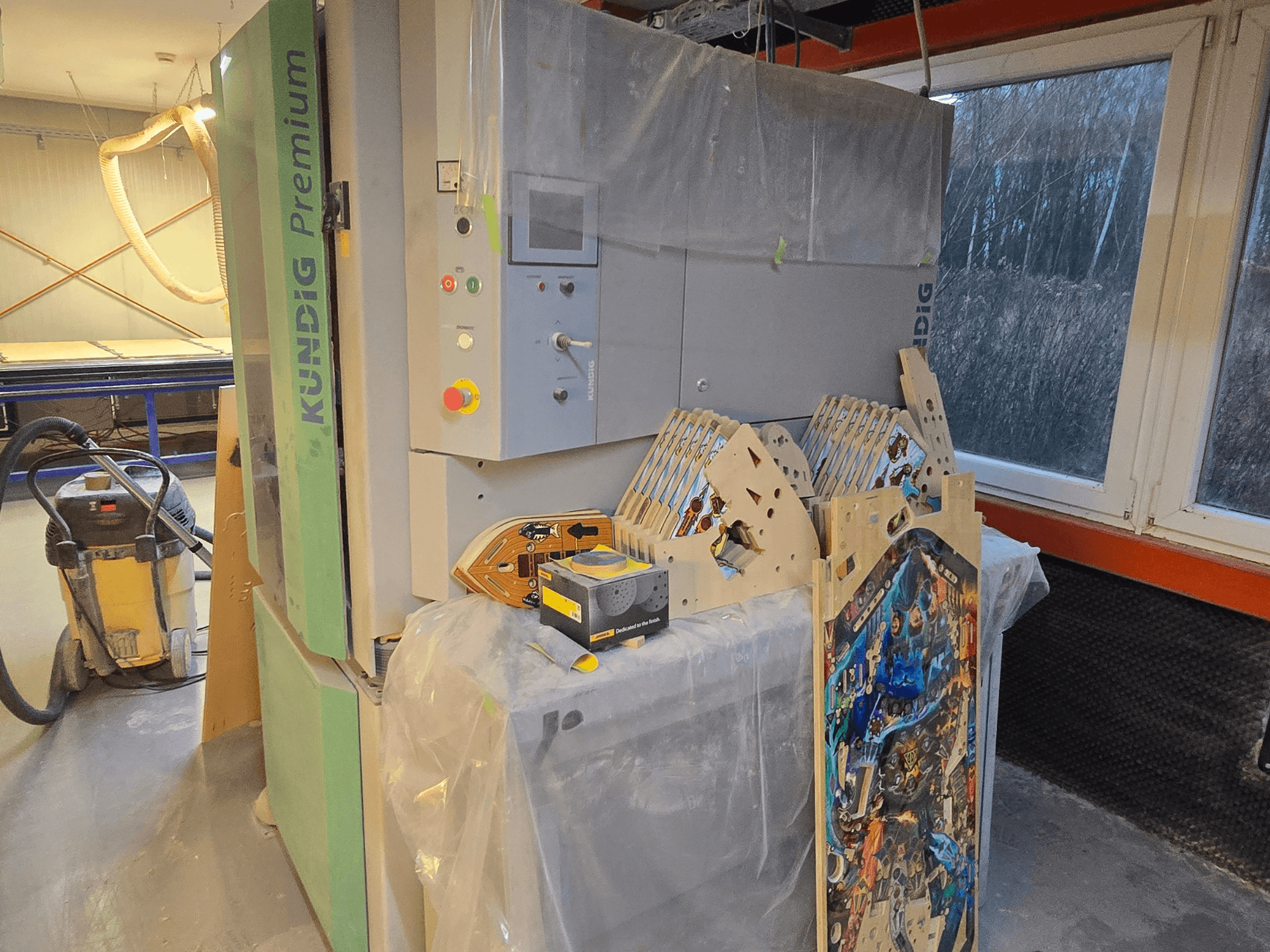 Kundig Premium-1 1100-C lintlihvpinker töökojas, näidatakse juhtpaneeli ja puidutükke, külgvaade, taustal tolmuimeja.