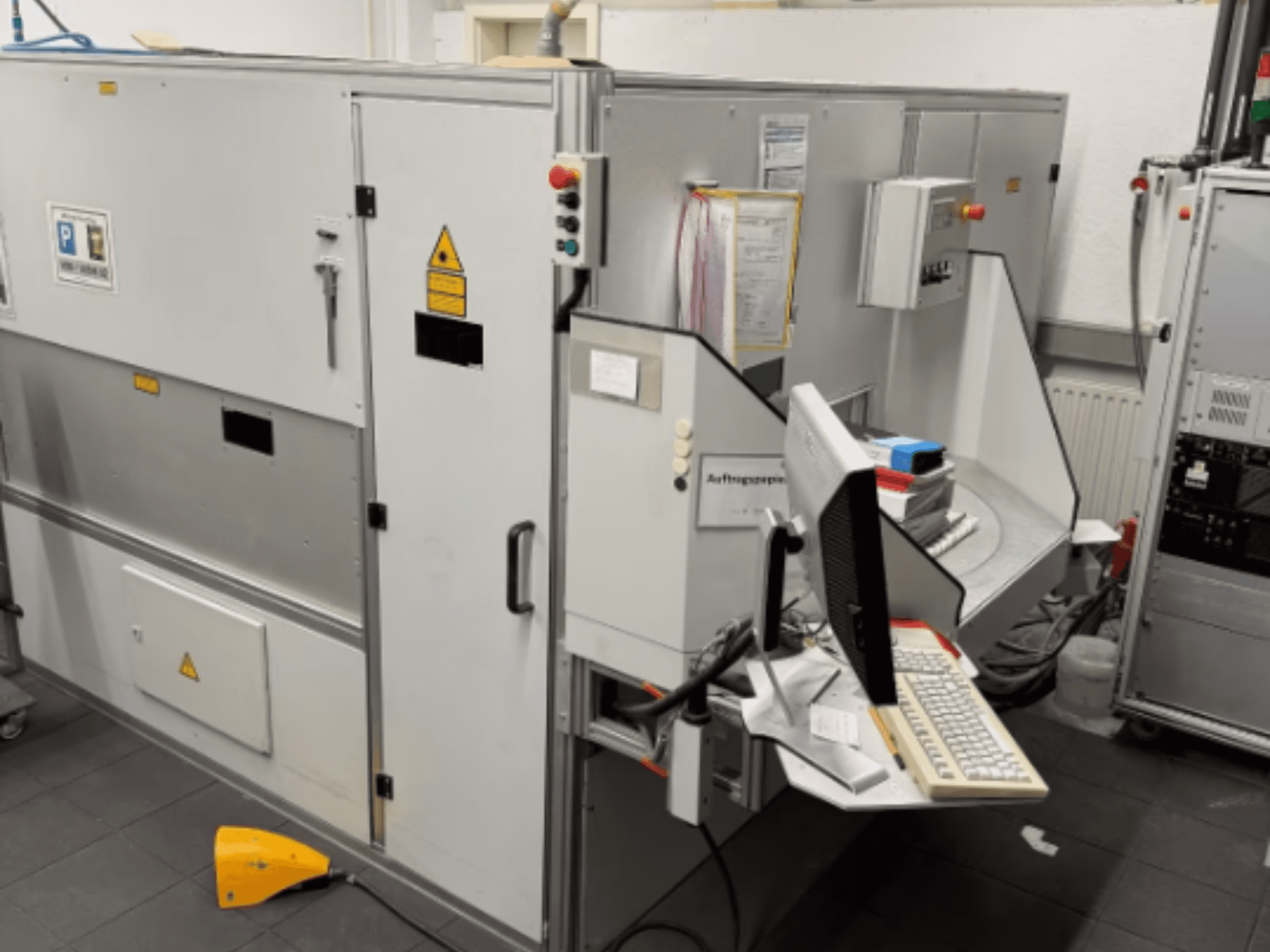 Tööstusmasin koos arvutiterminaliga, külgvaade; sisaldab juhtnuppe ja ohutushoiatusi.