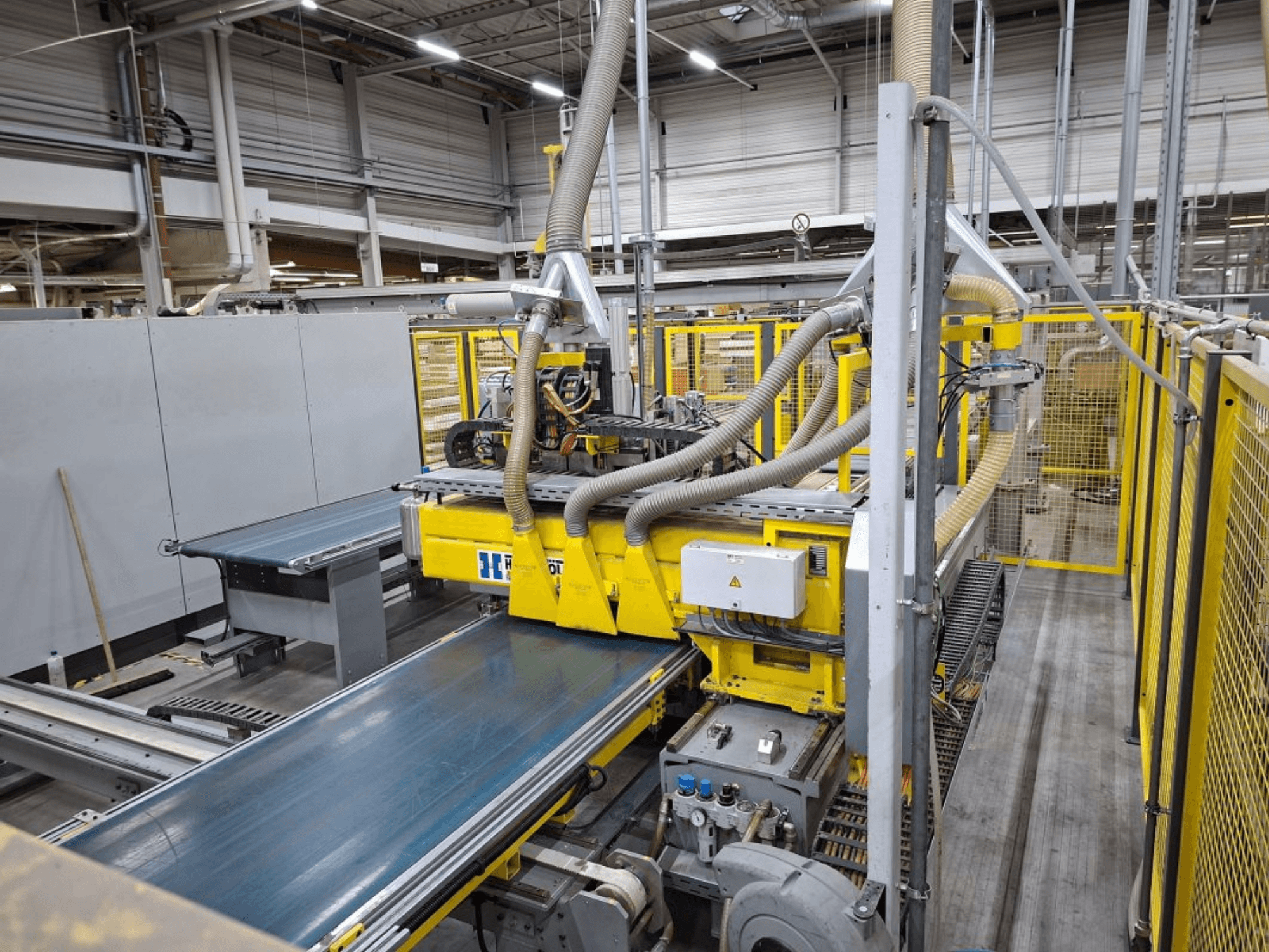 Tööstuslik konveierisüsteem külgvaates, millel on kollased masinad, juhtpaneel ja voolikukinnitused tootmiseks.
