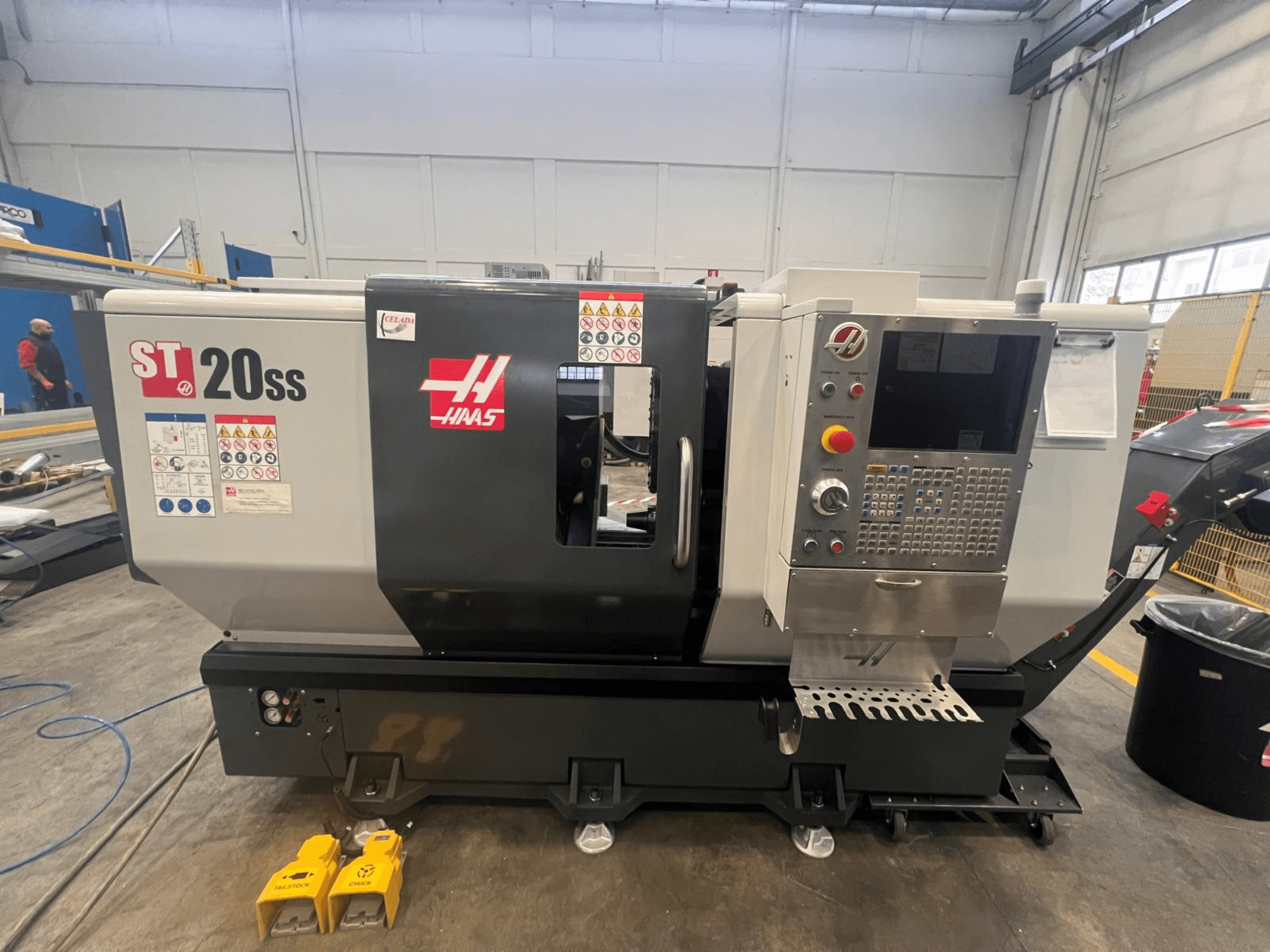 Haas ST 20SS CNC-teeninduspingi külgvaade, millel on juhtpaneel, ohutussildid ja jalgpedaalid põrandal.