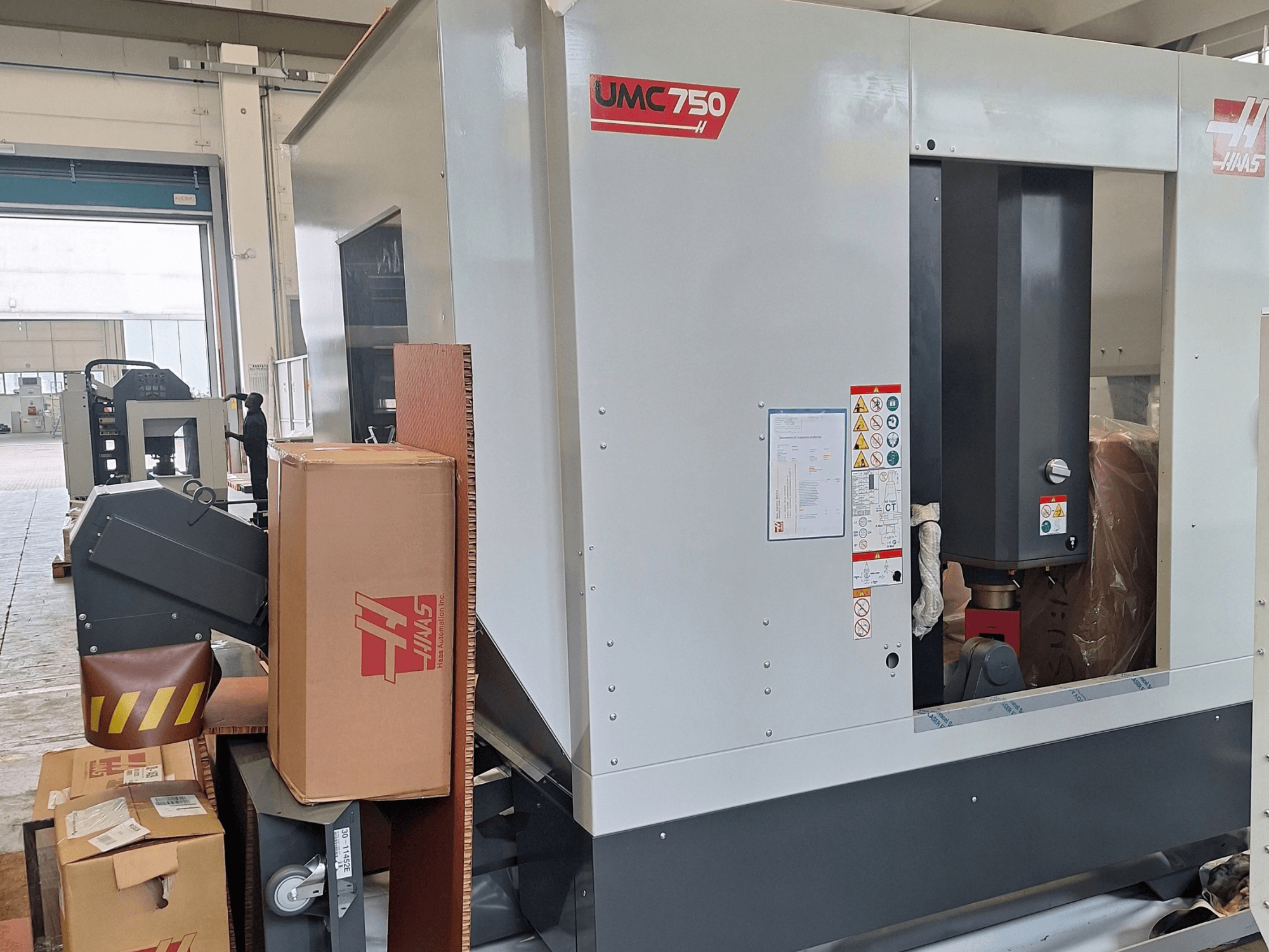 HAAS UMC-750 universaalne töötlemiskeskus, külgvaade, millel on näha juhtpaneel ja tööriistade hoiuruum koos hoiatussiltide ja pakenditega.