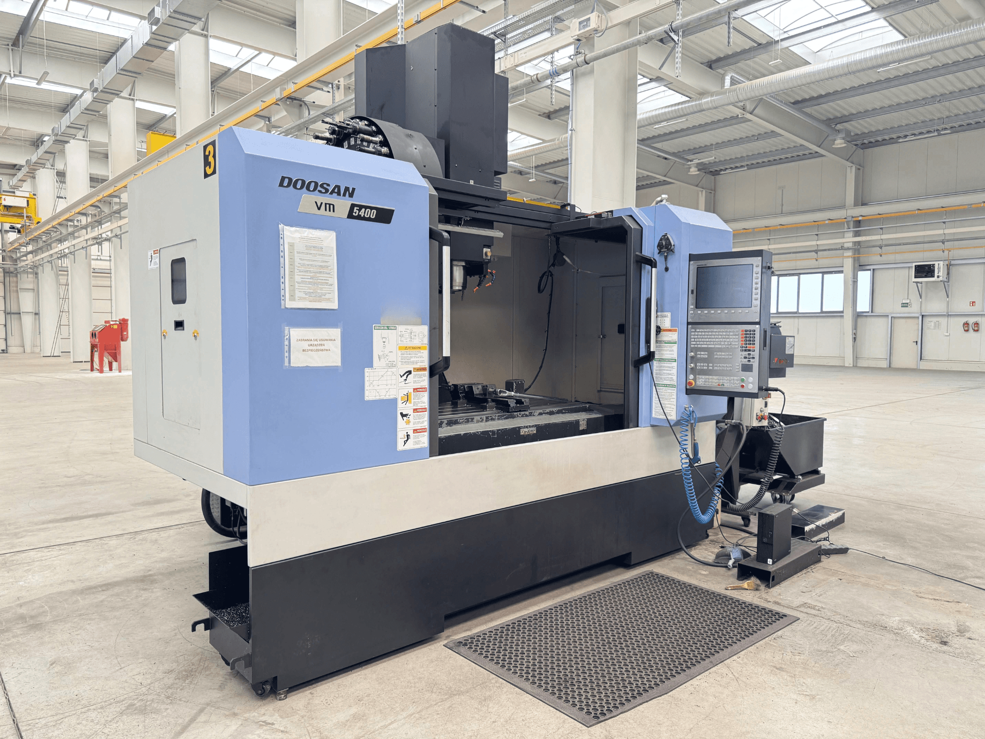 DOOSAN VM 5400 vertikaalne töötluskeskus, esiplaanil on juhtpaneel ja tööala avaras tööstuslikus keskkonnas.