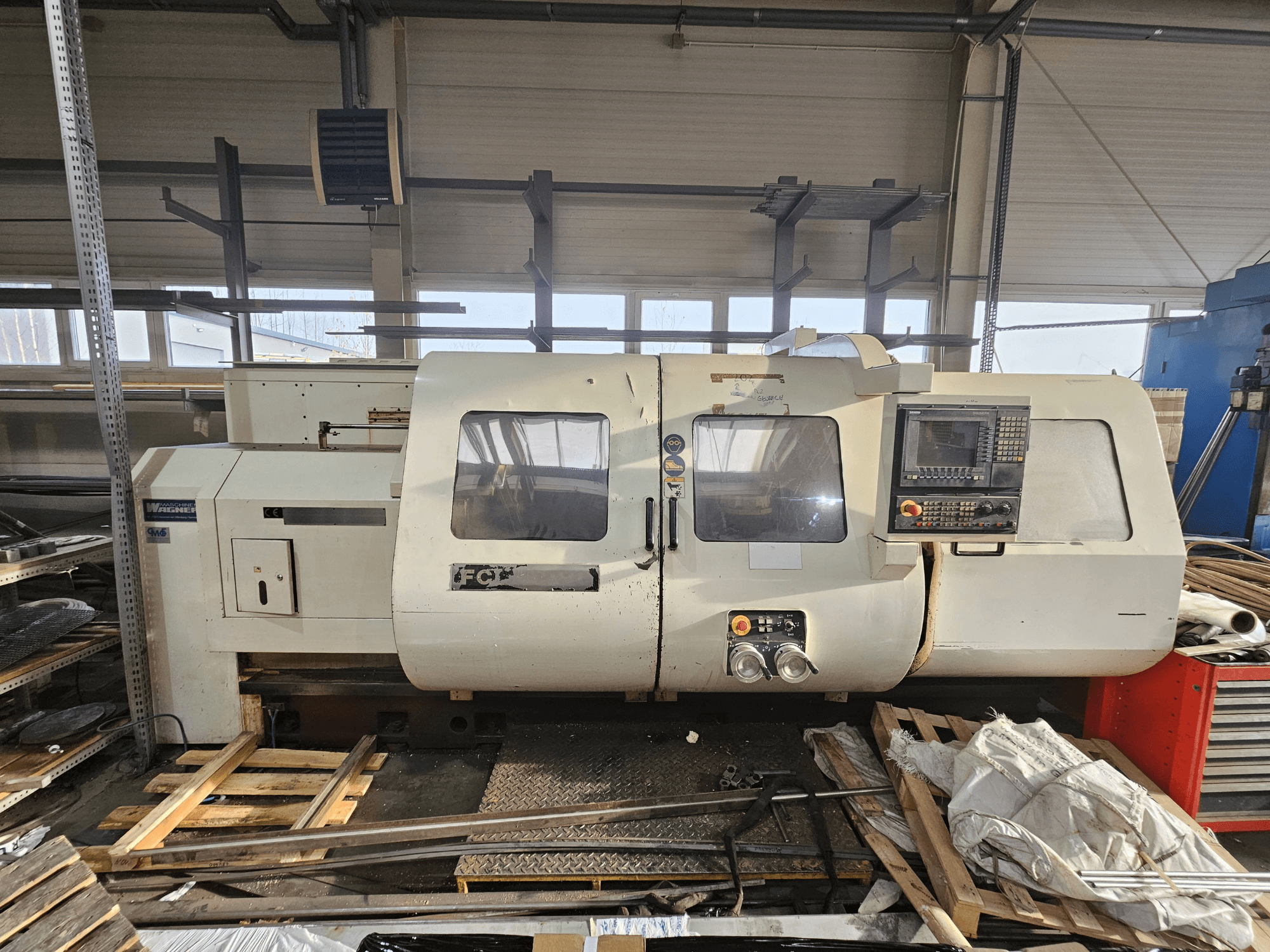 Wabeco CNC-tööpink, külgvaade, kus on juhtpaneel, aknad ja tööala; masinad puidust kaubaalustel töökojas.