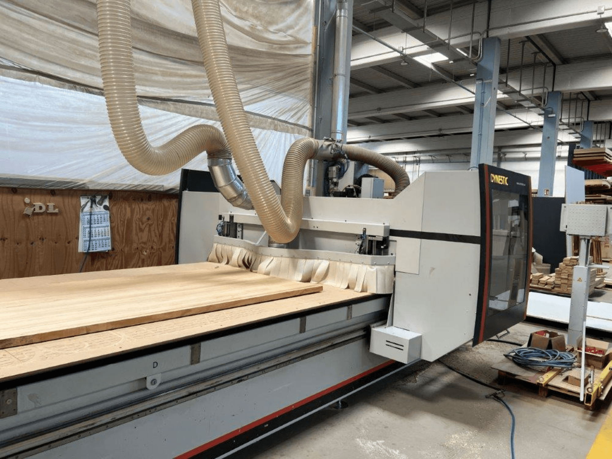 Dynestici CNC-tööpink, esiplaanil, millel on suur puidust toorik ja tolmueemaldusvoolik töökojas.