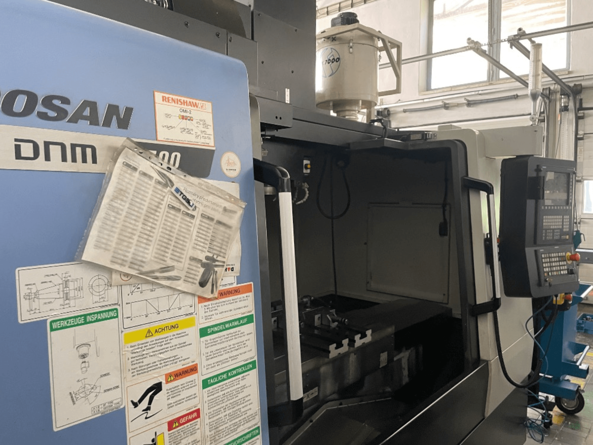 DOOSAN DNM 5700 vertikaalne töötluskeskus, esiplaanil on näidatud juhtpaneel, ohutussildid ja masina üksikasjad.