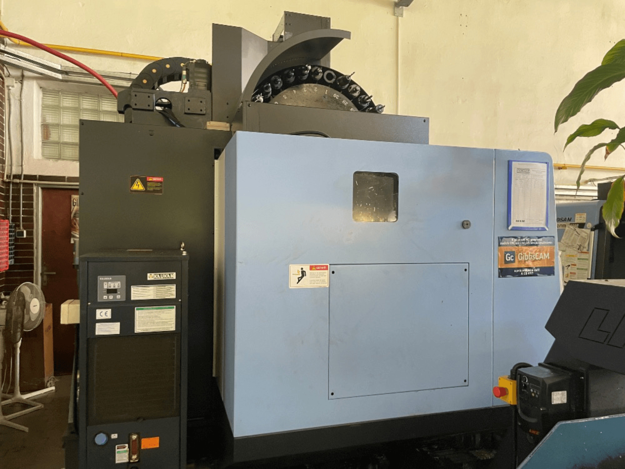 DOOSAN DNM 5700 vertikaaltöötluskeskuse esiplaan, kus on näidatud juhtpaneel, ohutussildid ja masina funktsioonid.