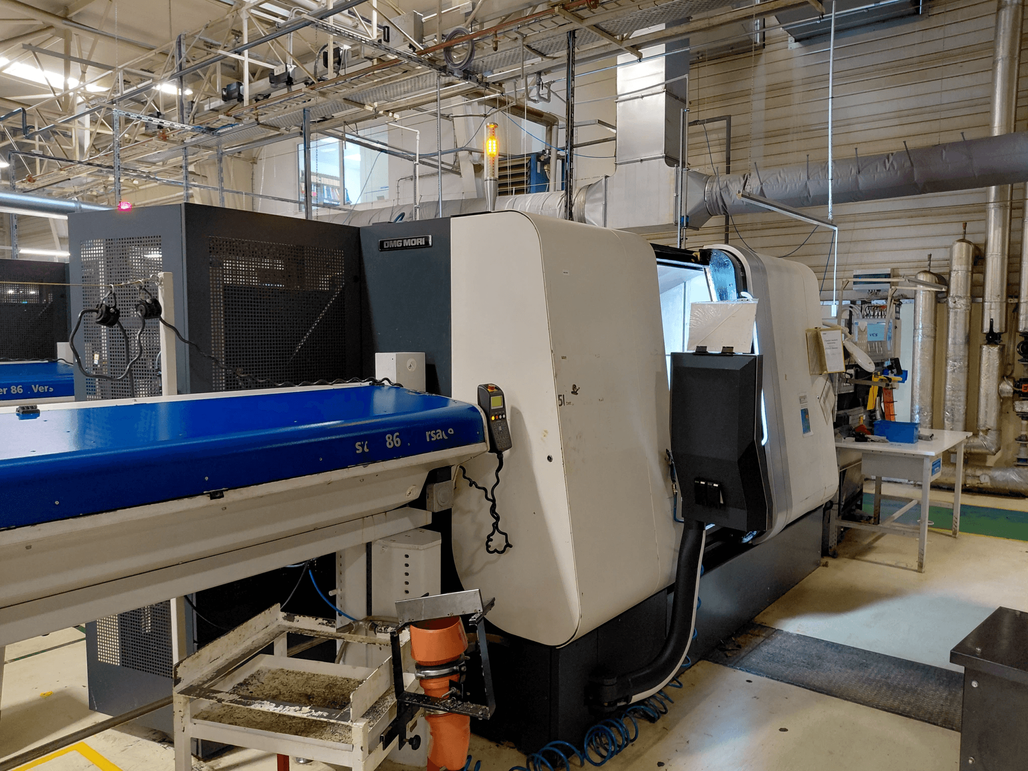 DMG MORI Sprint 65 lineaarne mitmepingeline treipink, külgvaade juhtpaneeli ja konveieriga, mis on nähtav töökojas.