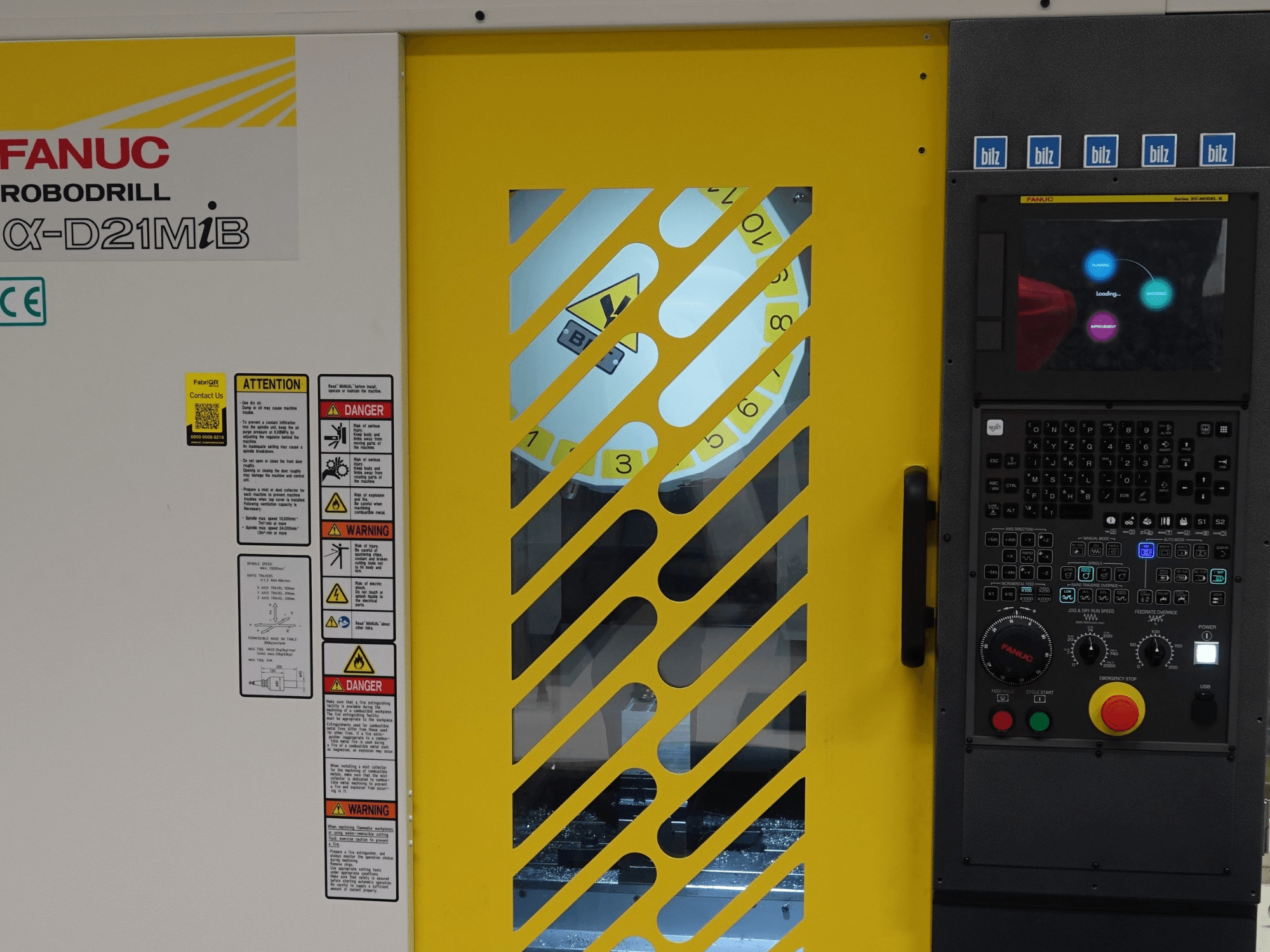 FANUC α-D21MiB vertikaalse töötlemiskeskuse esivaade, millel on näha ohutushoiatused, juhtpaneel ja kollane ohutusuks.