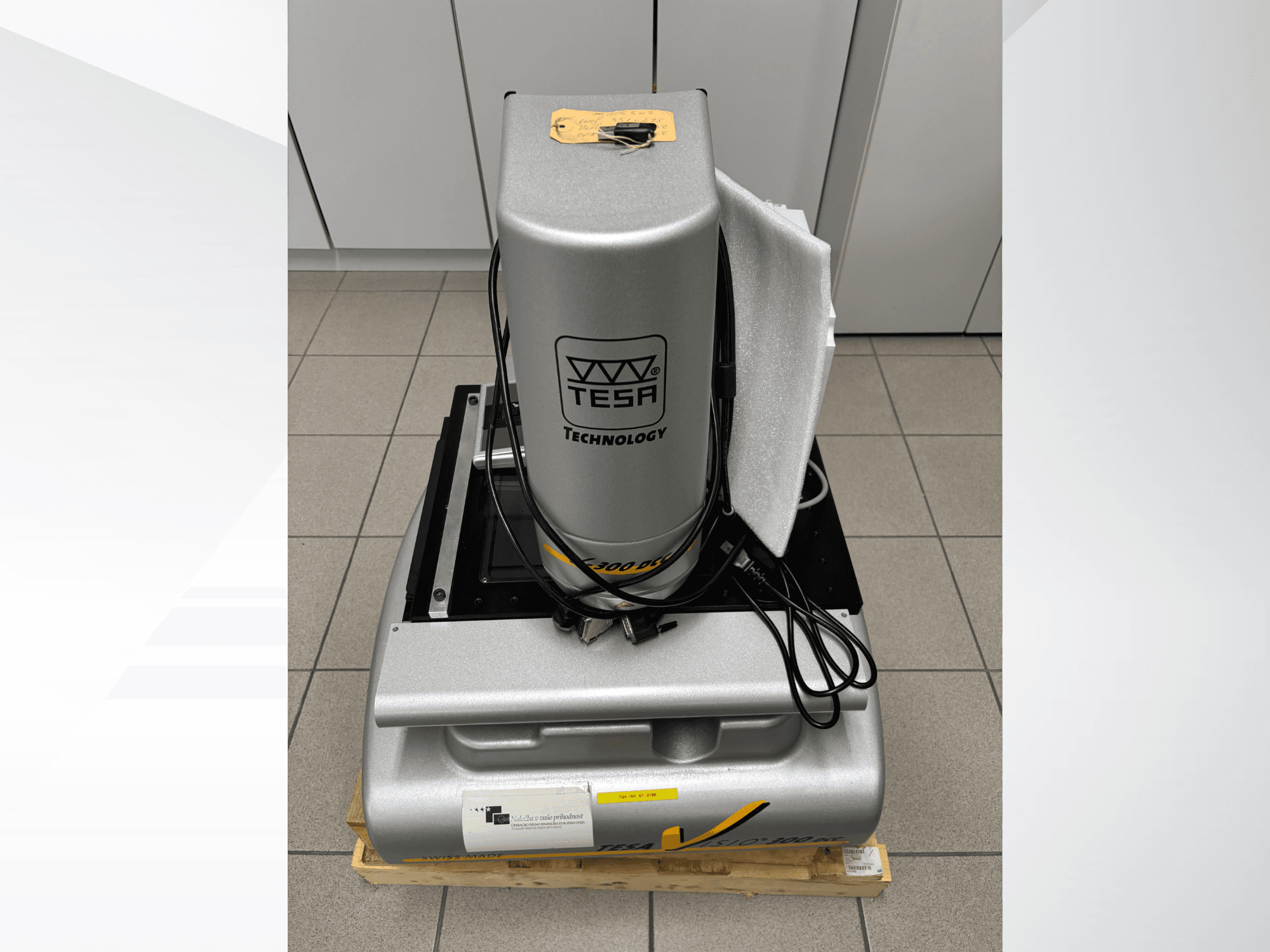 TESA Visio 300 DCC optiline mõõtmismasin esiplaanil koos kaablitega, puidust kaubaalusel ja nähtaval kaitsepakendis.