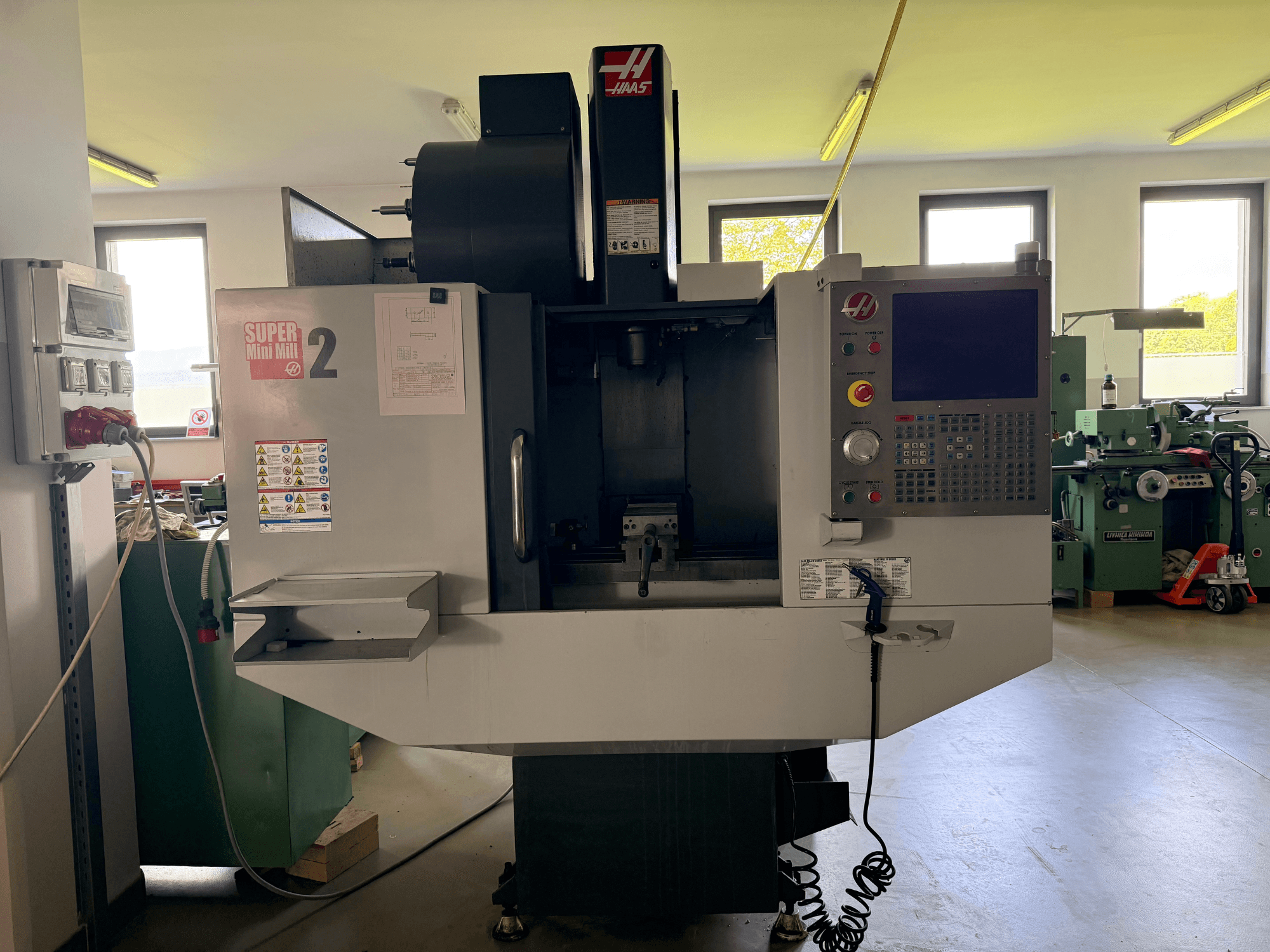 Haas Mini Mill 2 vertikaalne töötluskeskus esiplaanil, kus on näha juhtpaneel ja tööriistade ala koos hoiatussiltidega.