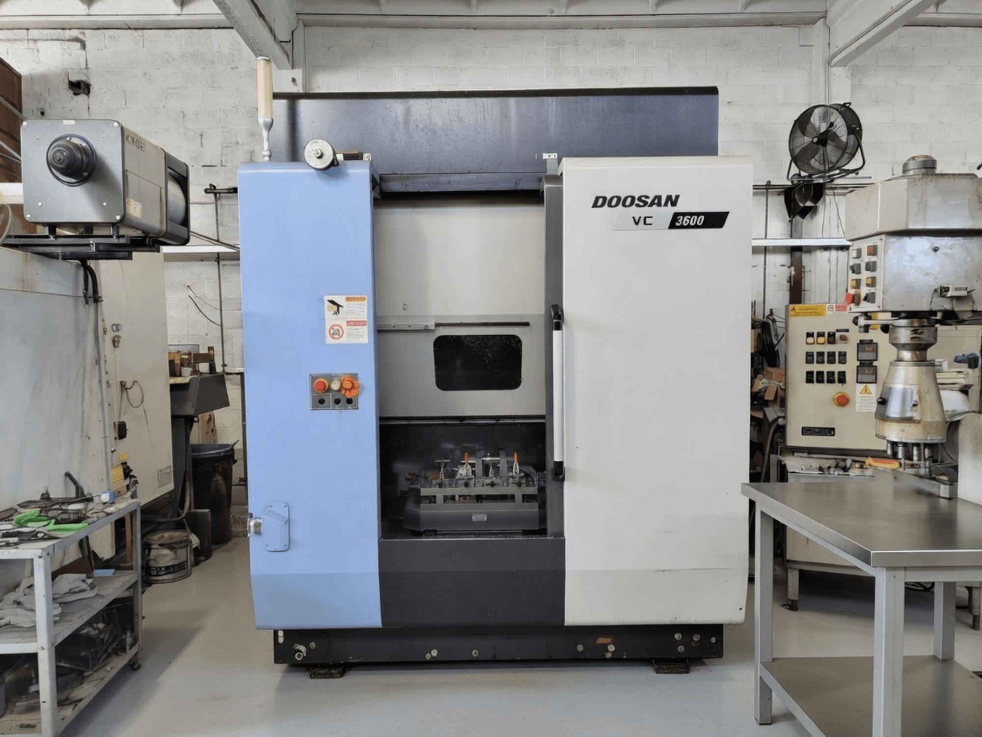 DOOSAN VC 3600 vertikaalne töötlemiskeskus esiplaanil, kus on juhtpaneel ja avatud uks, mis avab tööruumi.