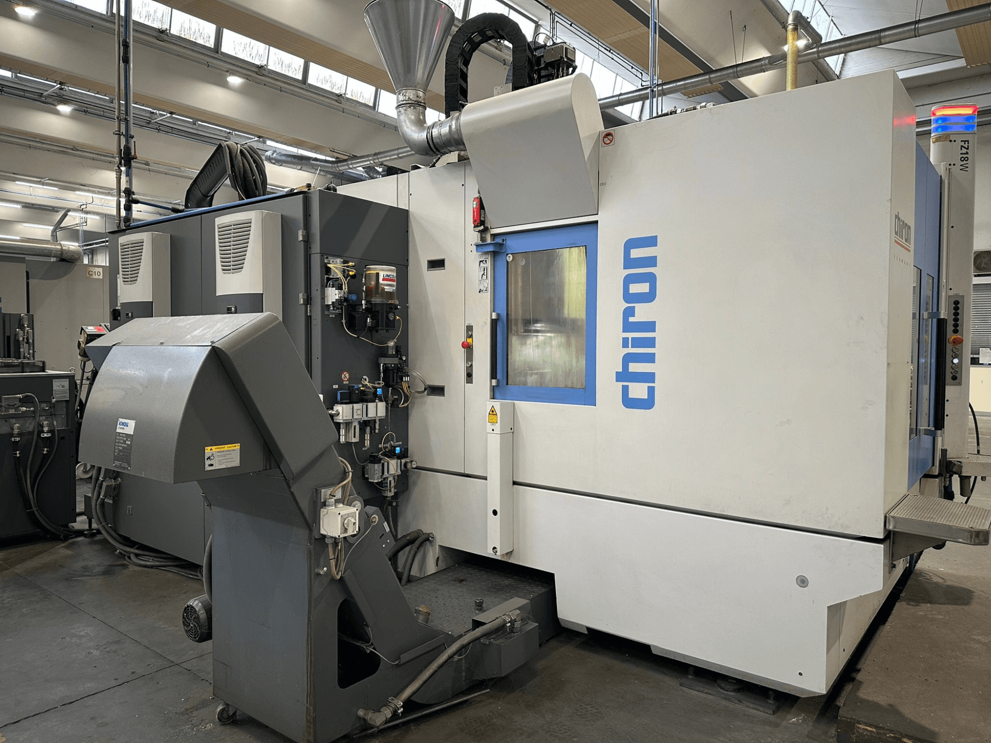 Chiron CNC-tööpink halli ja valge värviga, esiplaanil, millel on juhtimisseadmed, torud ja tolmueemalduskapuut.