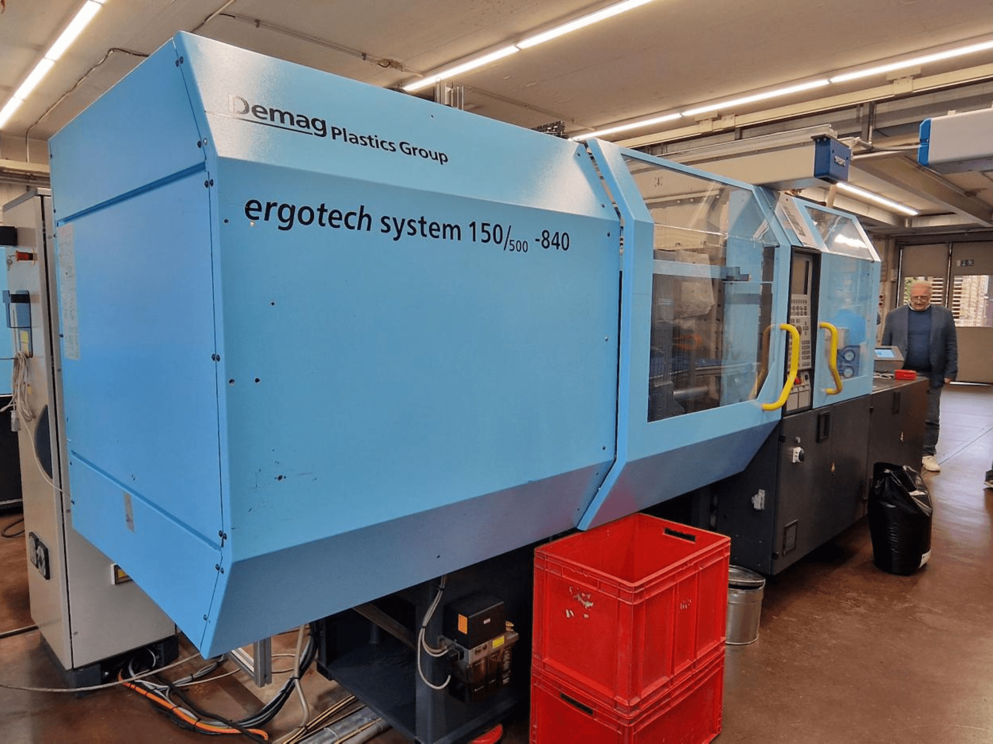 Demag Ergotech System 150/500-840 sinine survevalu masin, külgvaade, taustal kastid ja operaator.