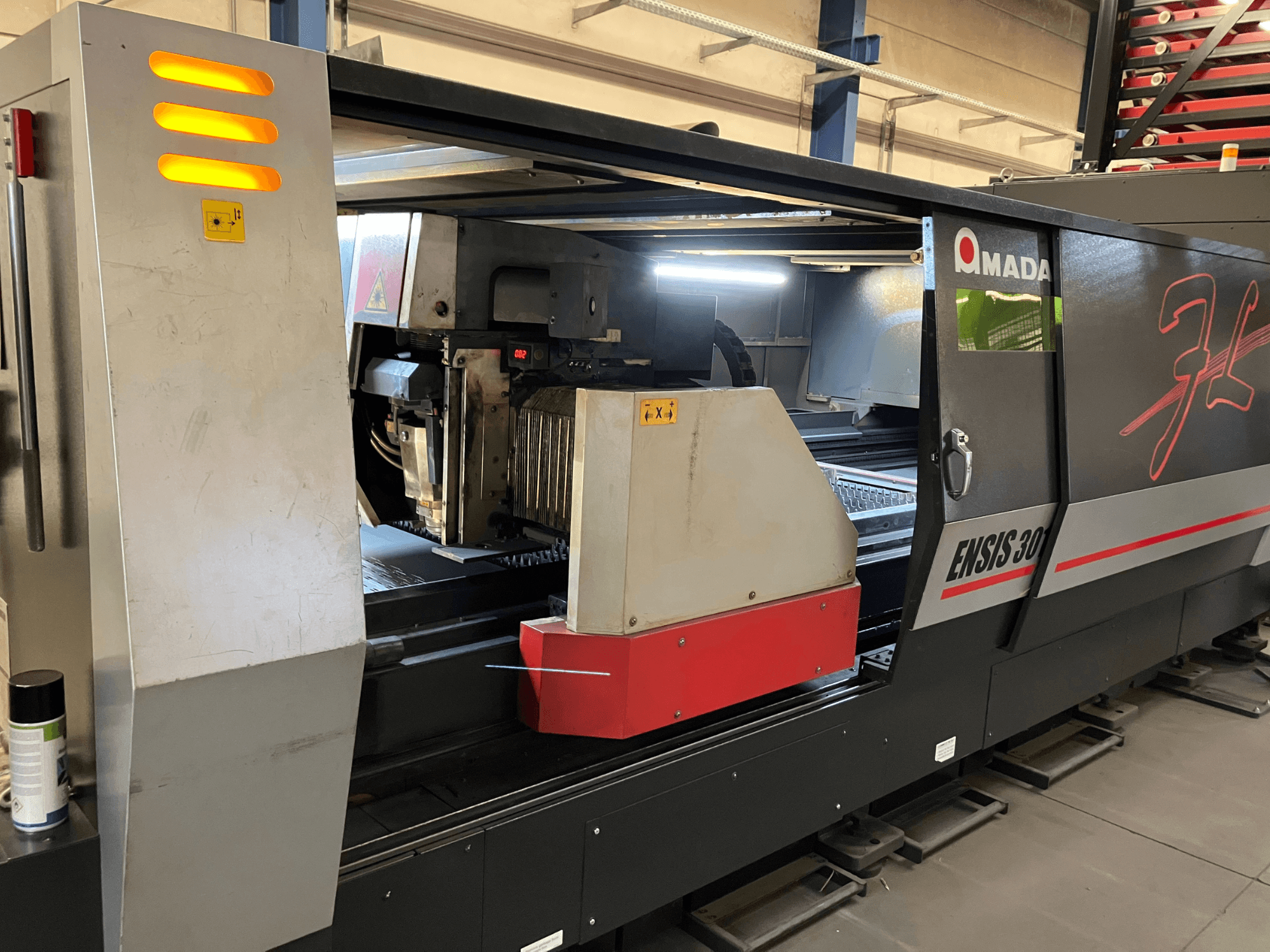 AMADA ENSIS 3015 AJ kiudoptiline laserlõikepink, esiplaanil, millel on kujutatud lõikamispea ja juhtimisseadmed metallist raami sees.