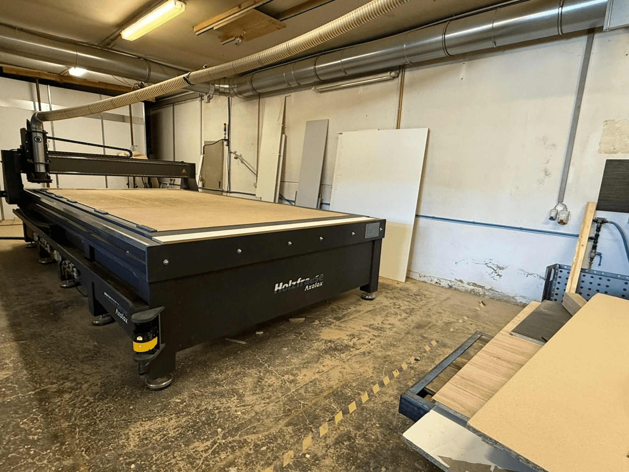 Holz-Her Axos 5 CNC-freesüsteem töökojas, esiplaanil töölaud, tolmueemaldussüsteem ja puitpaneelid lähedal.