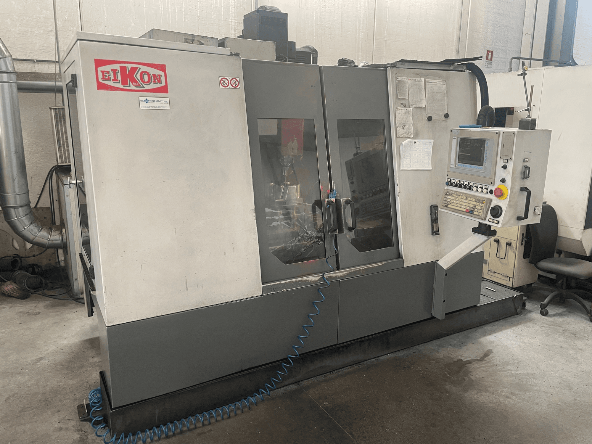 EIKON CNC-tööpink ettepoole vaadatuna, millel on juhtpaneel, suured klaasuksed ja sinine õhuvoolik põrandal.