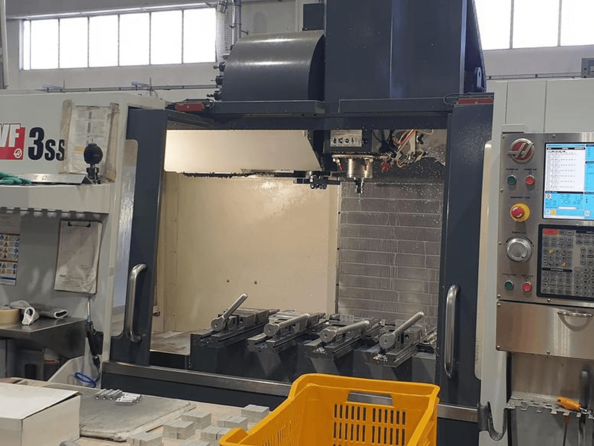 HAAS VF-3SS vertikaalne töötlemiskeskus, esiplaanil CNC juhtpaneel, tööriistad ja oranž korv.