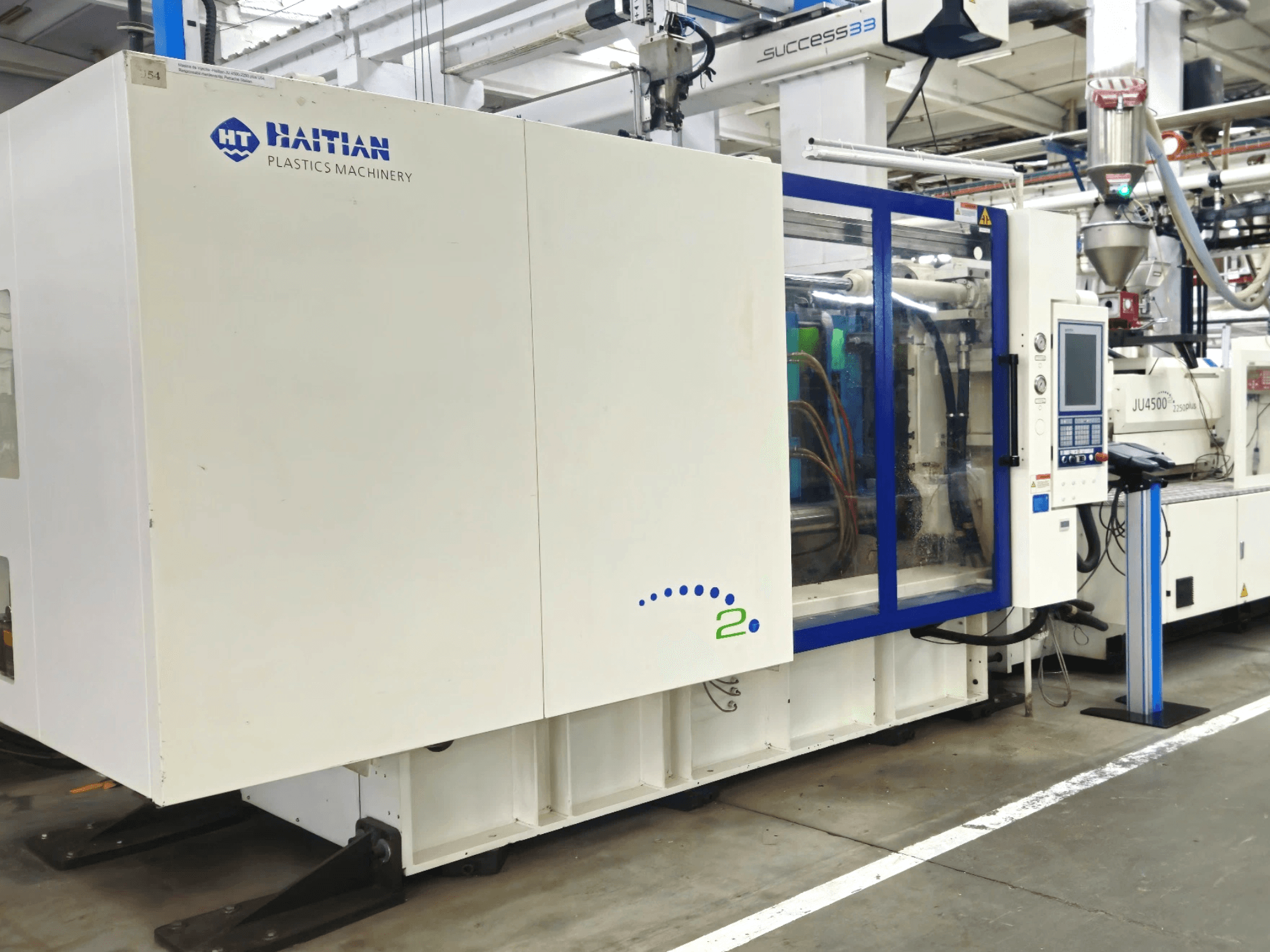 Haitian Plastics Machinery mudel, esiplaanil, mis näitab masinaid koos juhtpaneeli ja nähtavate torudega tööstuslikus keskkonnas.