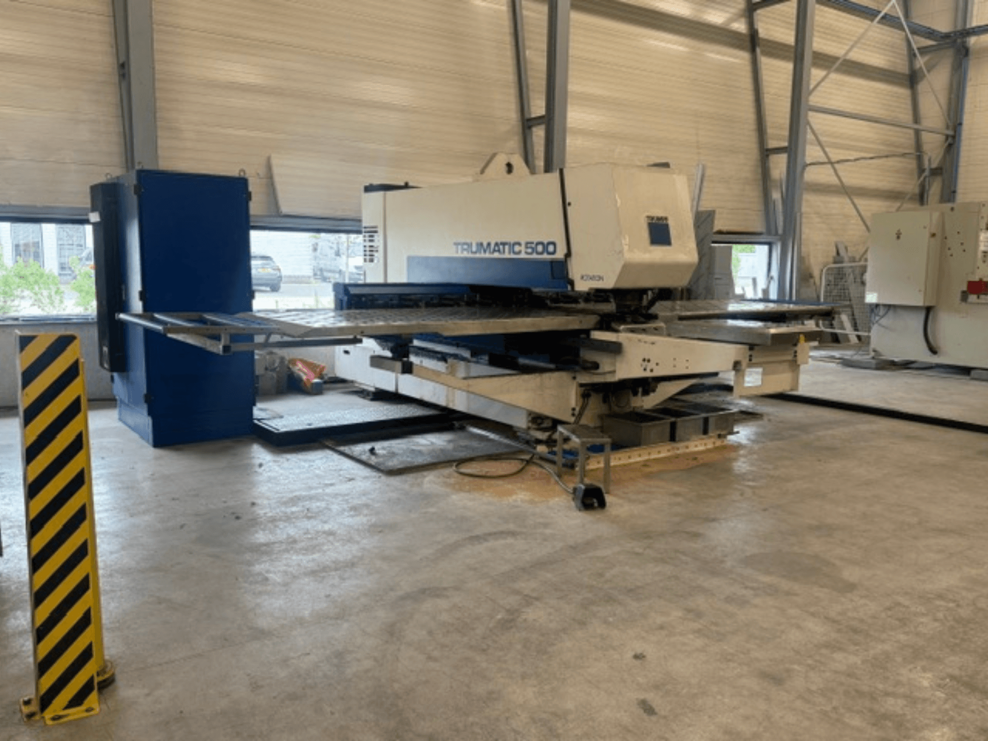 Töökojas asuv CNC-stantsimismasin TRUMPF Trumatic 500, külgvaade, millel on näha masin ja kollaste triipudega ohutuspost.