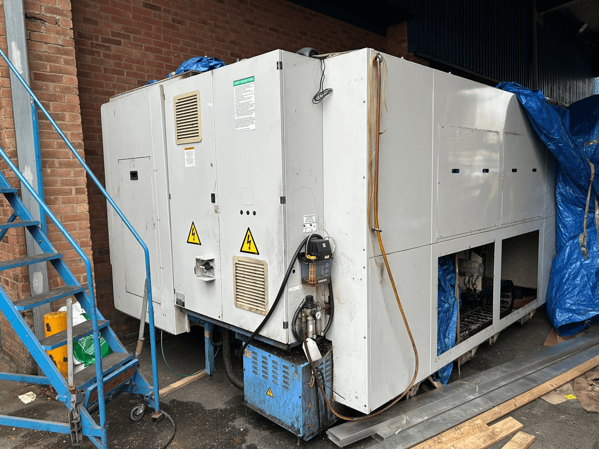 Suure tööstusliku jahutusseadme külgvaade, kaubamärk ja mudel ebaselge, millel on näha elektriühendused, hoiatusmärgid ja sinine tarpkate.