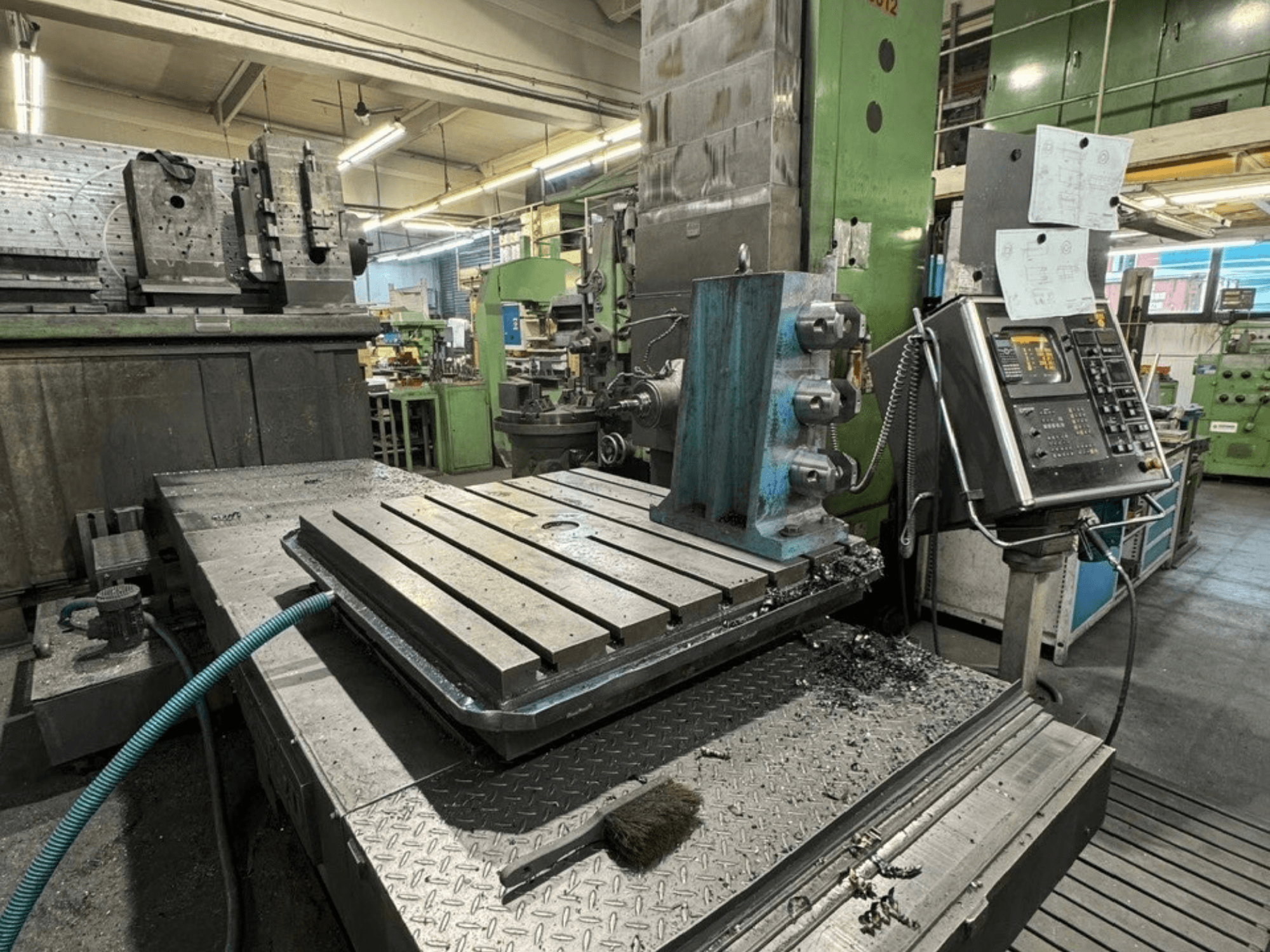 Töökojas asuv puurmasin JUARISTI 12/TS1.7, külgvaade, millel on näha soontega metalllaud ja selle kõrval asuv juhtpaneel.