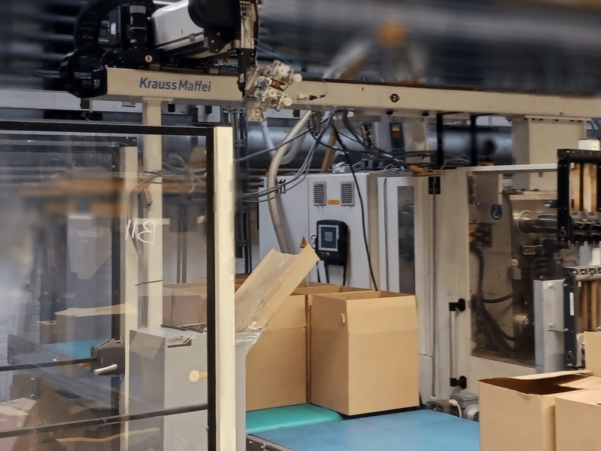 Krauss Maffei automatiseerimissüsteem tehases, esiplaanil kastid konveieril ja masinaosad töös.