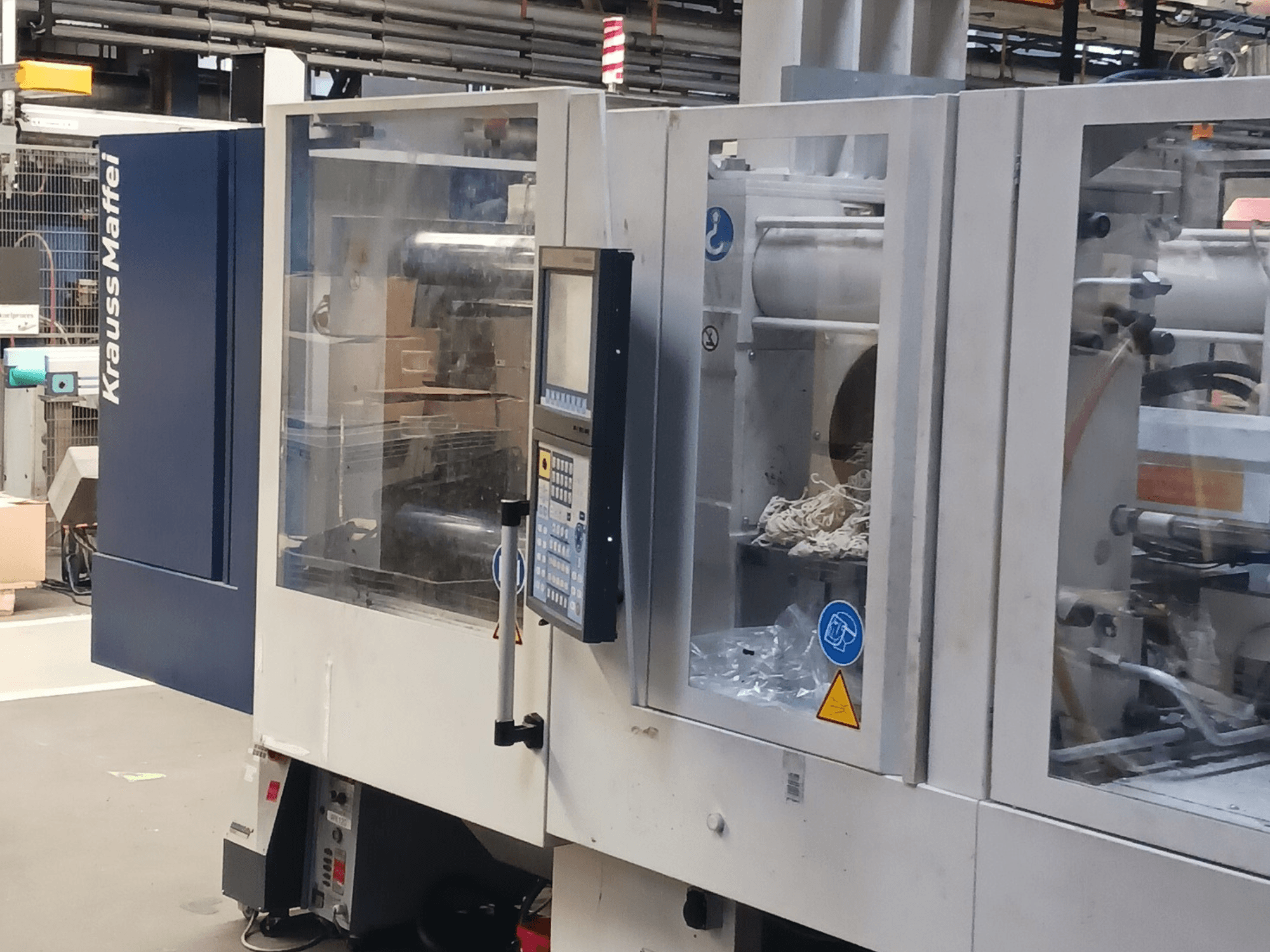 KraussMaffei survevalu masin, külgvaade, kus on näidatud juhtpaneel, masinad ja pakendimaterjalid taustal.